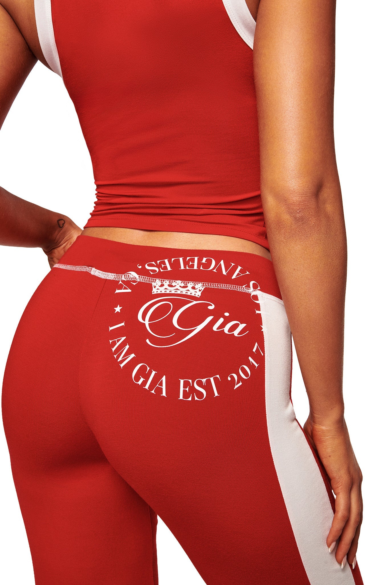 A person wears the I.AM.GIA BLARE PANELLED TRACKPANT in red, a straight leg athletic pant with a white circular "Gia, I AM GIA EST 2017, LOS ANGELES" print on the back.