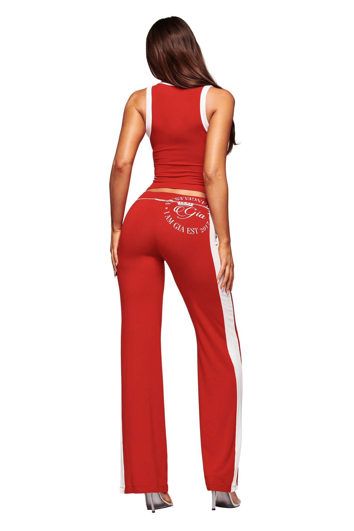 A woman wears the I.AM.GIA BLARE PANELLED TRACKPANT - RED, featuring white side stripes and back printed text, paired stylishly with high heels.