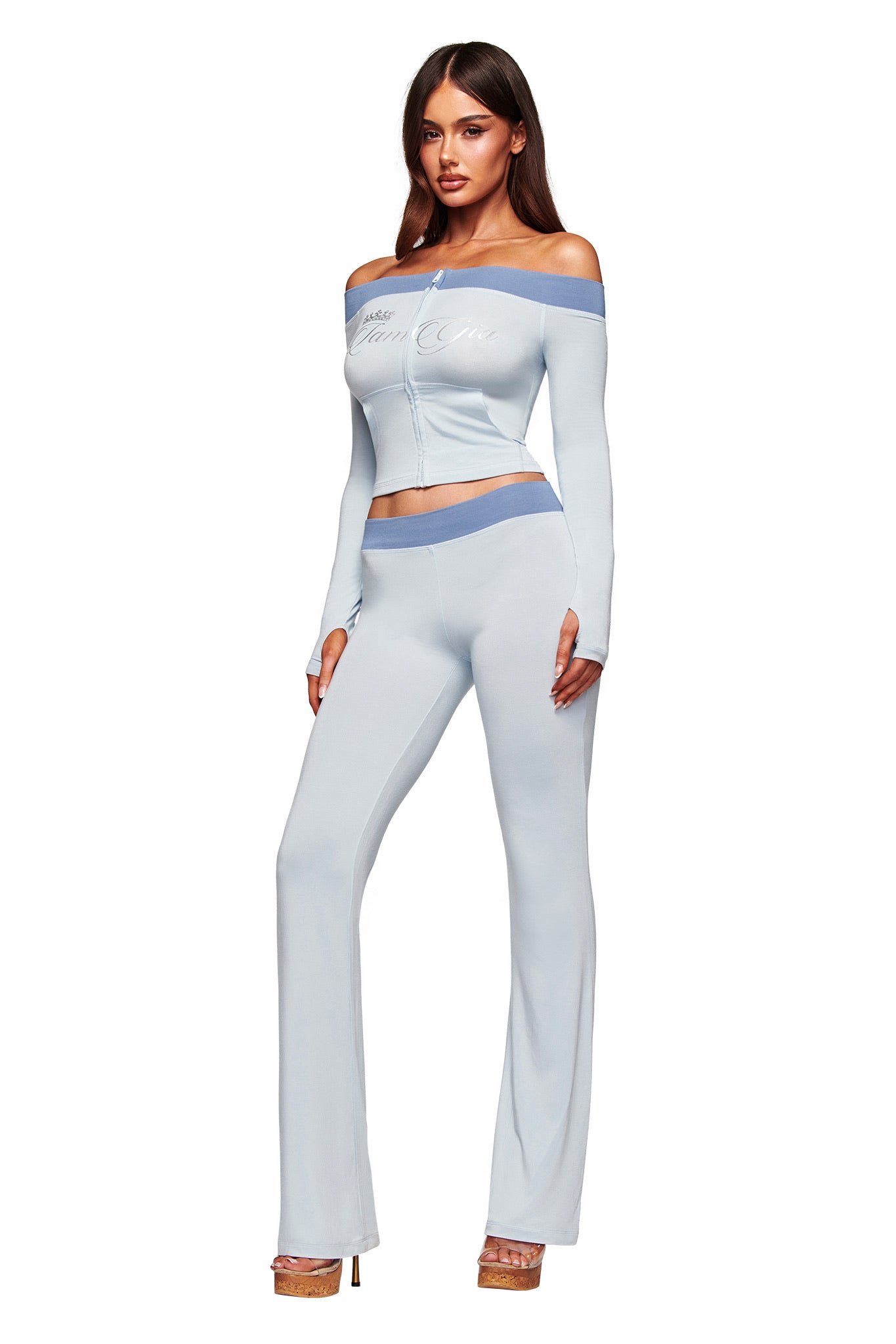 A woman models I.AM.GIA’s BLARE LOWER RISE TRACKPANT in two tone powder blue with a matching off-shoulder top, both in bamboo viscose, creating a fitted casual look. She pairs the outfit with beige high heels against a white background.