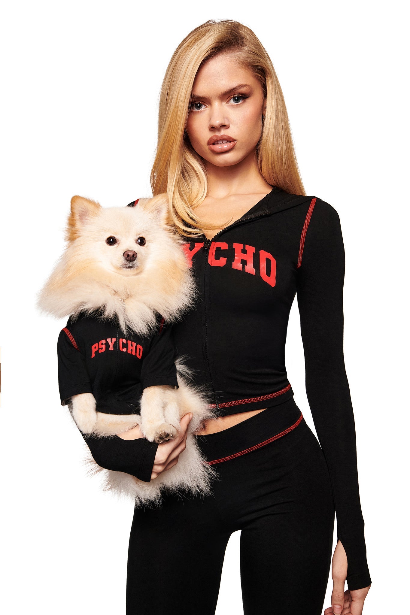 A woman in a black I.AM.GIA outfit reading "PSYCHO" holds her dog, who wears the matching I.AM.GIA BLARE BEST FRIEND DOG HOODIE - BLACK PSYCHO. Both face the camera confidently against a white background.