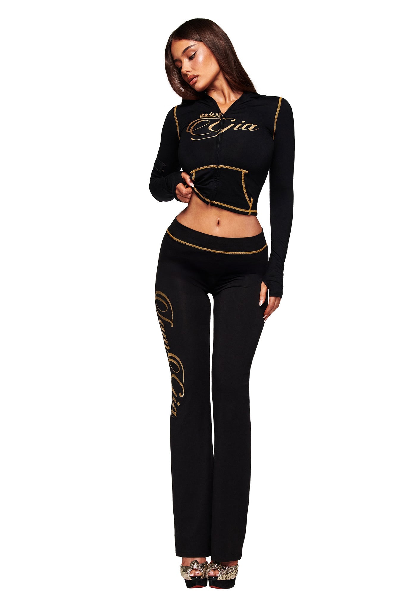 A woman models the I.AM.GIA BLARE FORM MULTI POCKET HOODIE in black and gold with cursive gold lettering, styled with a cropped top, flared pants, and high heels against a white background.