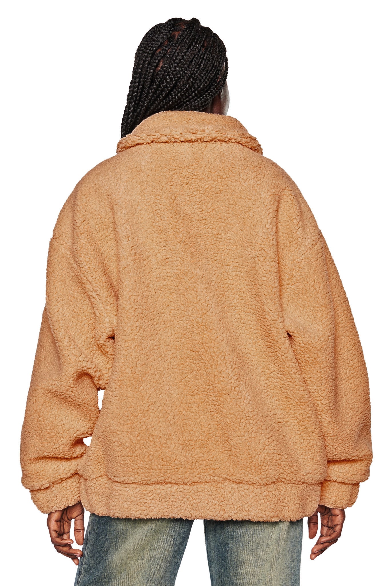 A person with braided hair is shown from the back, wearing a cozy, oversized PIXIE COAT in caramel from I.AM.GIA and blue jeans. The fluffy coat envelops the person's torso and arms completely.