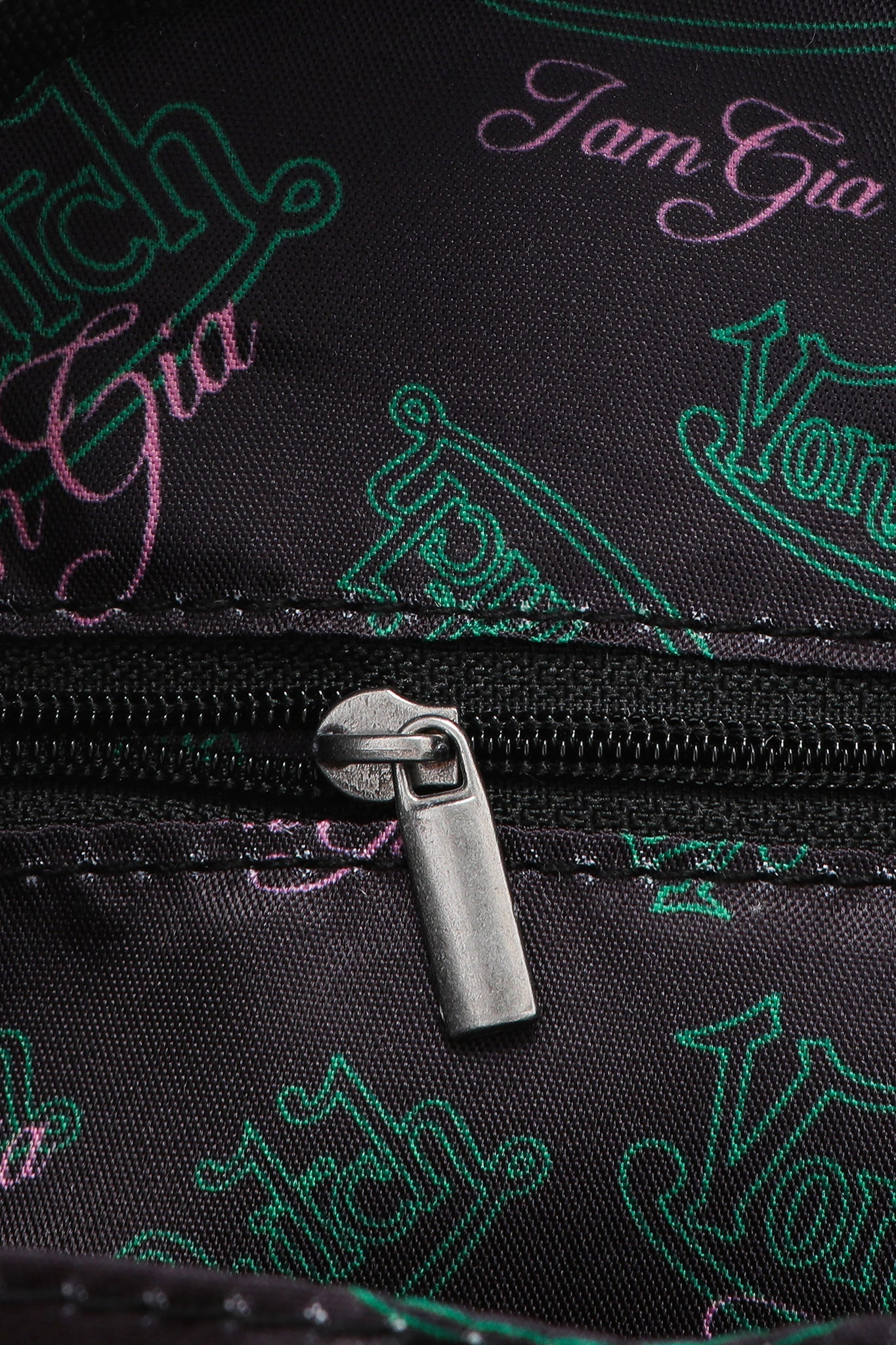 Close-up of the metallic zipper on black fabric with green and pink "Von Dutch" and "I.AM.GIA" logos highlights the I.AM.GIA X VON DUTCH MINI BOWLING BAG - LEOPARD, inspired by the iconic Von Dutch bowling bag.