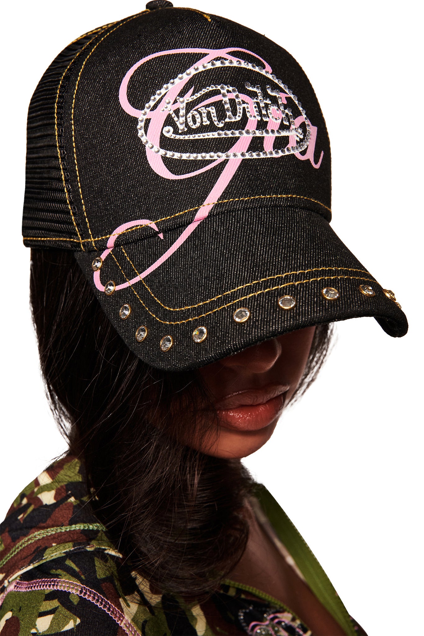 A person with long dark hair wears the I.AM.GIA X VON DUTCH TRUCKER HAT in indigo denim, featuring a diamante pink logo, partially covering their face. They wear a camouflage-patterned top against a white background.