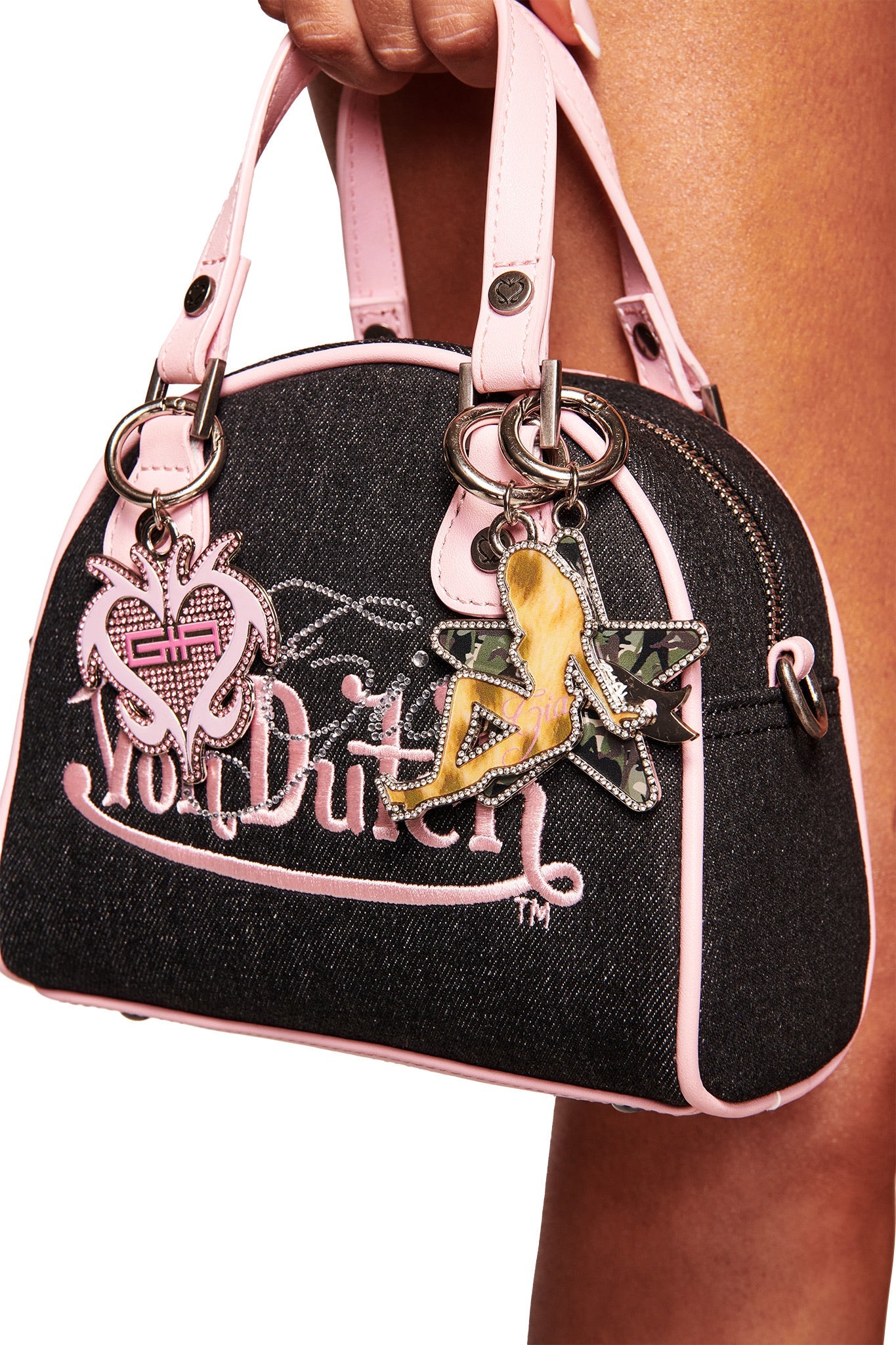 A hand holds the I.AM.GIA X VON DUTCH BOWLING BAG in indigo denim, featuring pink trim and handles, and decorated with a heart-shaped keychain and a silhouette woman keychain.