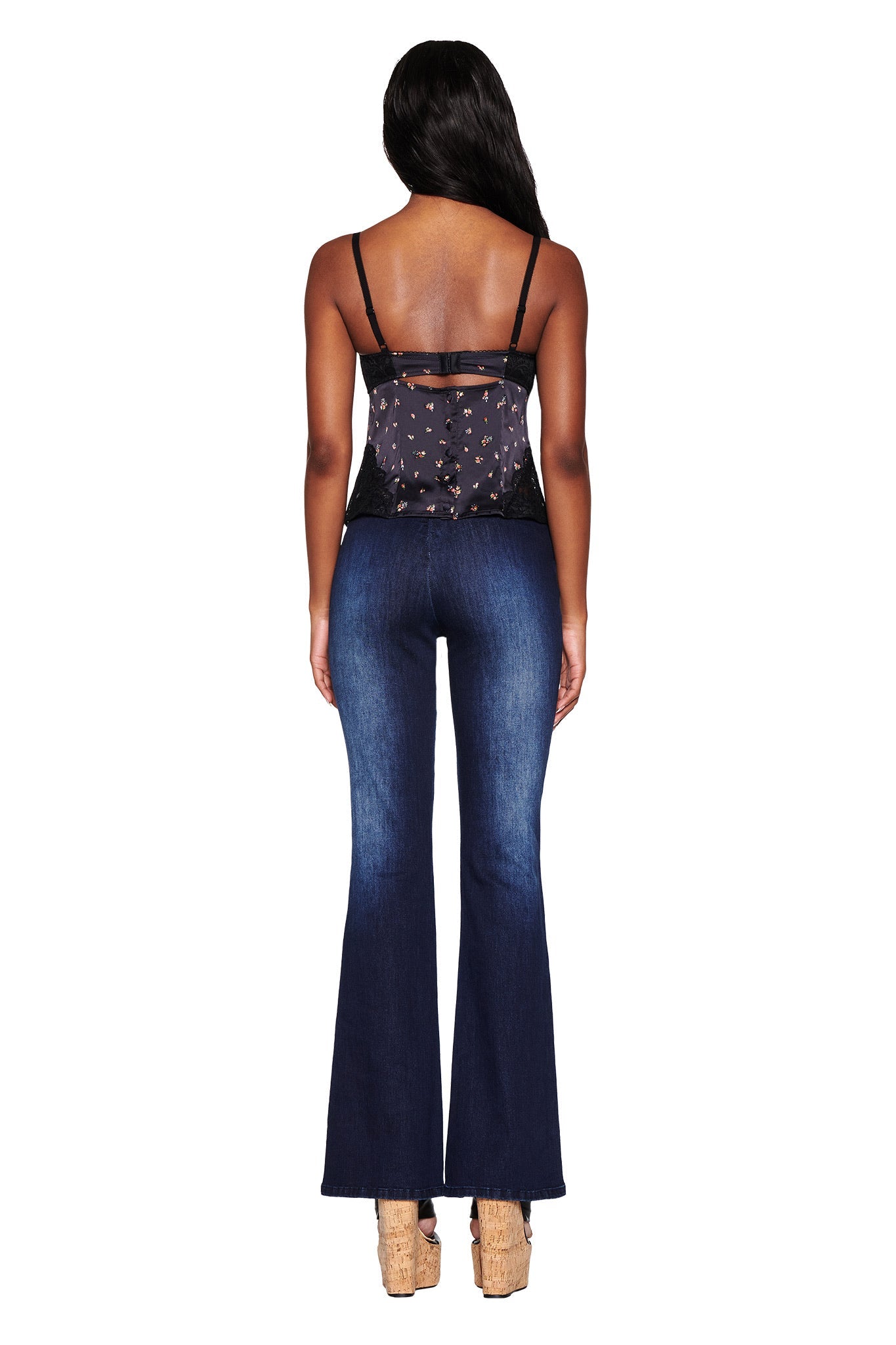 Rear view of a person with long hair, wearing an I.AM.GIA LUELLA CAMI TOP - BLACK : DITSY FLORAL and blue flared jeans, paired with brown wedge sandals against a plain white background.