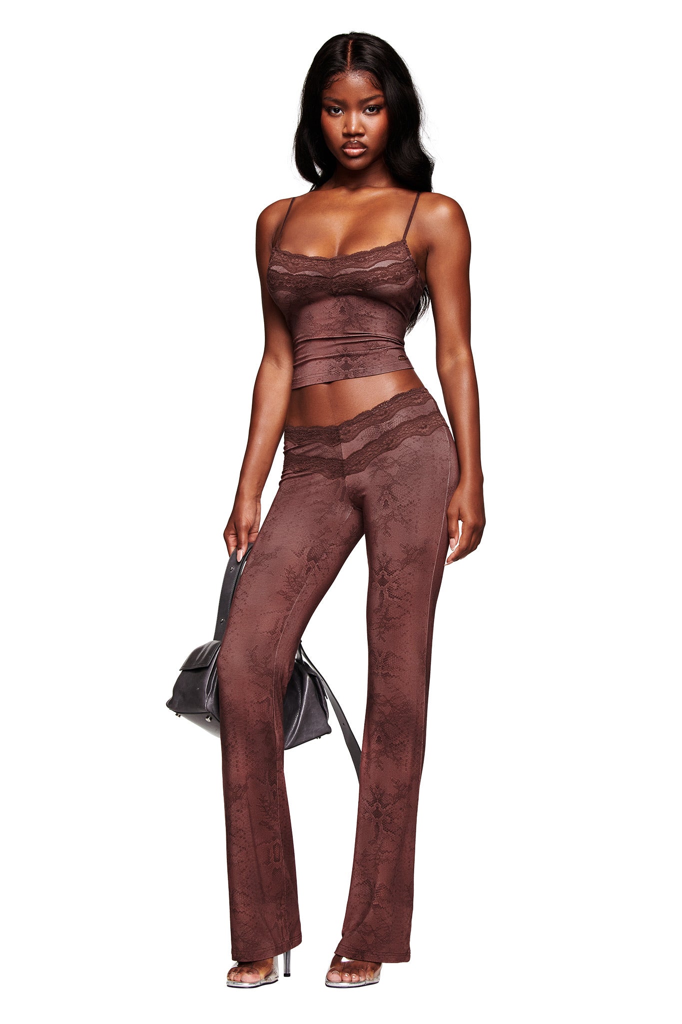 A woman wearing the I.AM.GIA BEAU PANT in brown with a matching lace-patterned crop top and holding a black handbag stands confidently against a white background.