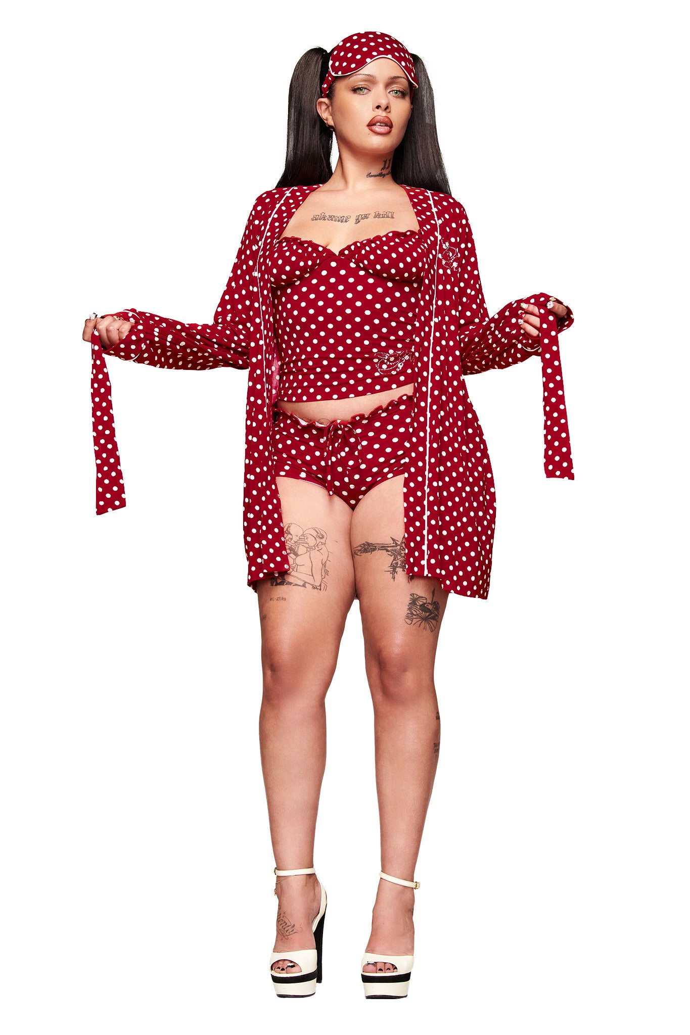 A woman in the MELANIE SLEEPWEAR - RED POLKA DOT set by I.AM.GIA, featuring a matching robe and headband, stands confidently with long dark pigtails, visible tattoos, and white heels against a white background.