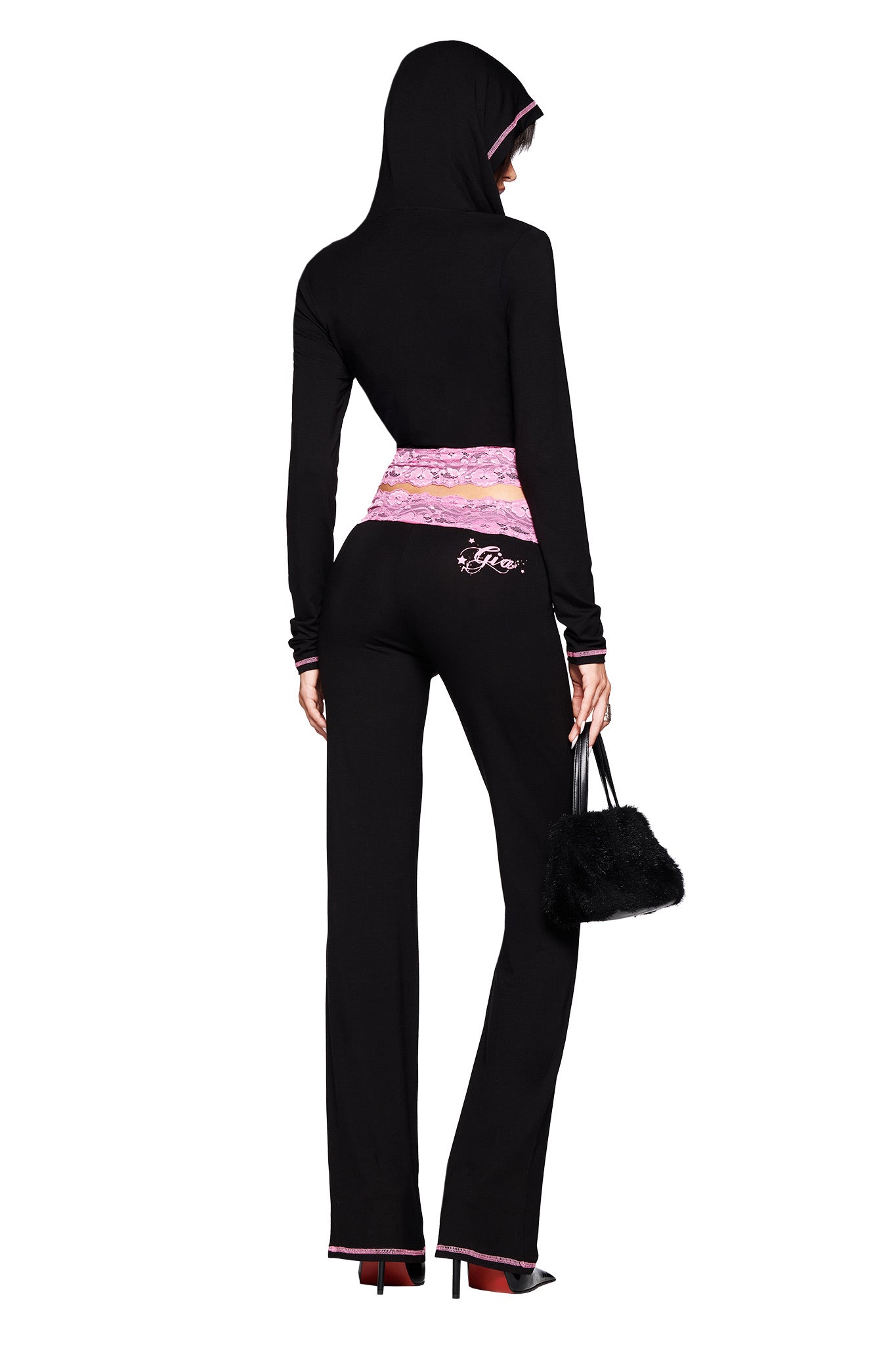 A person in the I.AM.GIA TARSHA PANT - BLACK/PINK, featuring a mid rise waist and pink lace waistband, holds a small black fuzzy handbag and stands with their back to the camera, highlighting the slim leg silhouette.
