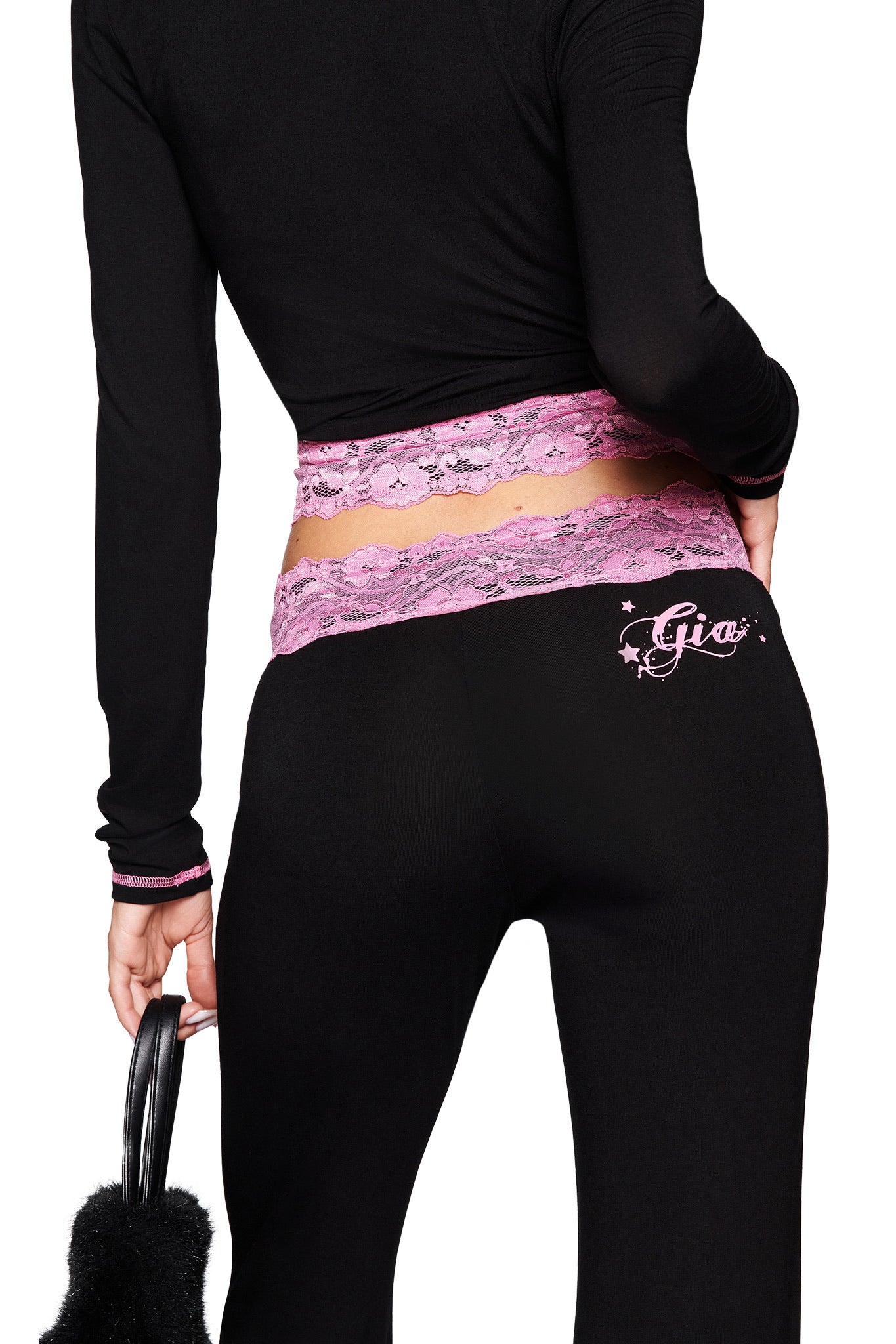 A person is seen from behind in I.AM.GIA’s TARSHA PANT - BLACK/PINK, featuring a pink lace mid rise waist and "Gia" on the back, paired with a matching black long-sleeve top and holding a black fuzzy accessory.