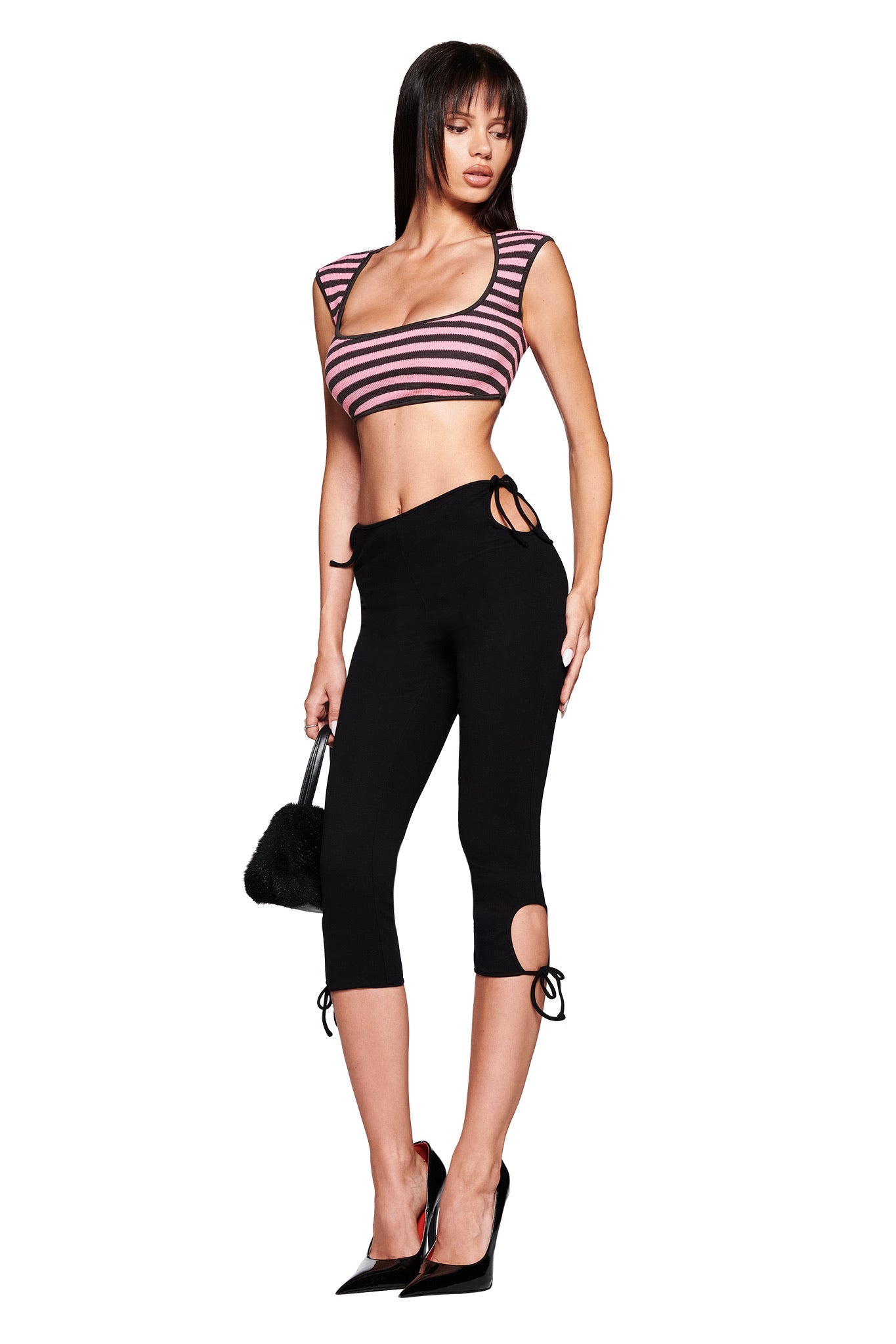 A woman poses against a white background wearing I.AM.GIA's YUNA CAPRI - BLACK, paired with a striped crop top, black high heels, and a small black handbag.