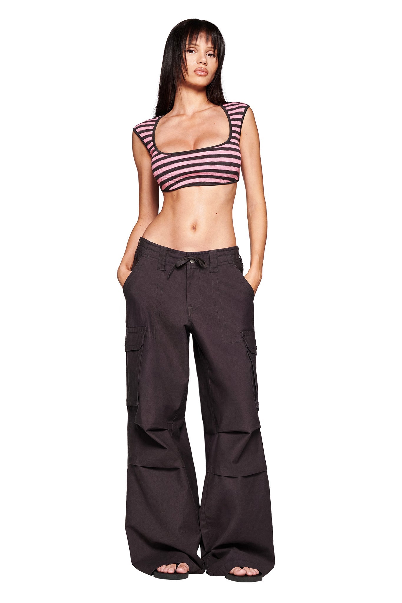 A woman stands barefoot against a white background, wearing the I.AM.GIA JAXXON PANT in grey with a baggy leg silhouette and a pink and black striped crop top, hands in her pockets.