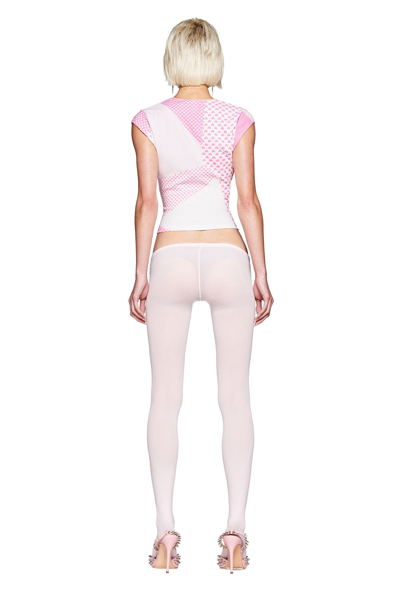 A person with short blond hair faces away, wearing the I.AM.GIA FAFI TOP - PINK featuring a Gia X Fafi X Faline graphic, paired with sheer white tights and pink feather-detail high-heeled sandals against a white background.