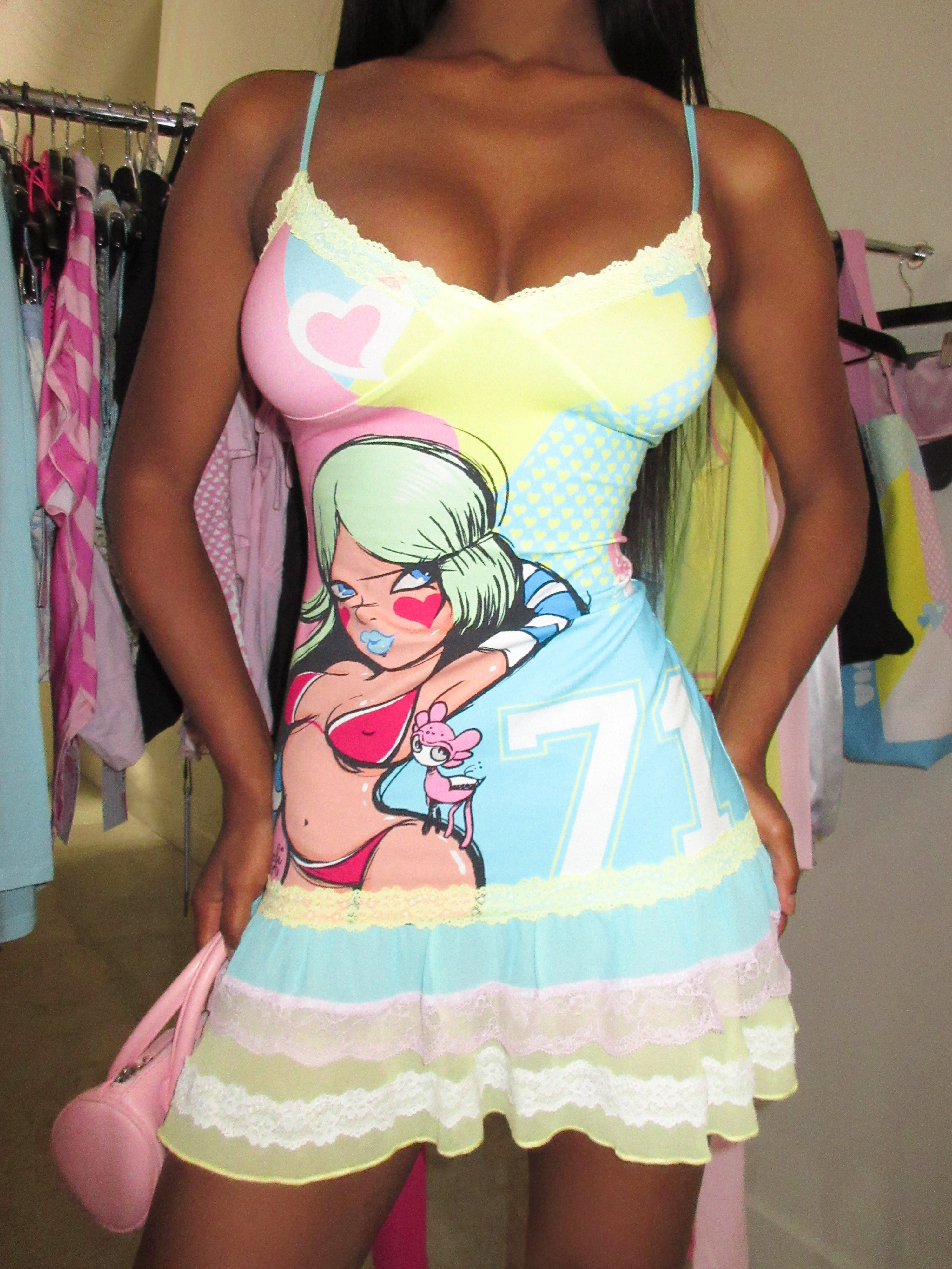 A woman wears the I.AM.GIA FAFI MINI DRESS - BLUE, a pastel stretch jersey cami mini dress with spaghetti straps and a cartoon girl graphic, hearts, and numbers from the FAFI collaboration. She holds a small pink purse with clothing racks behind her.