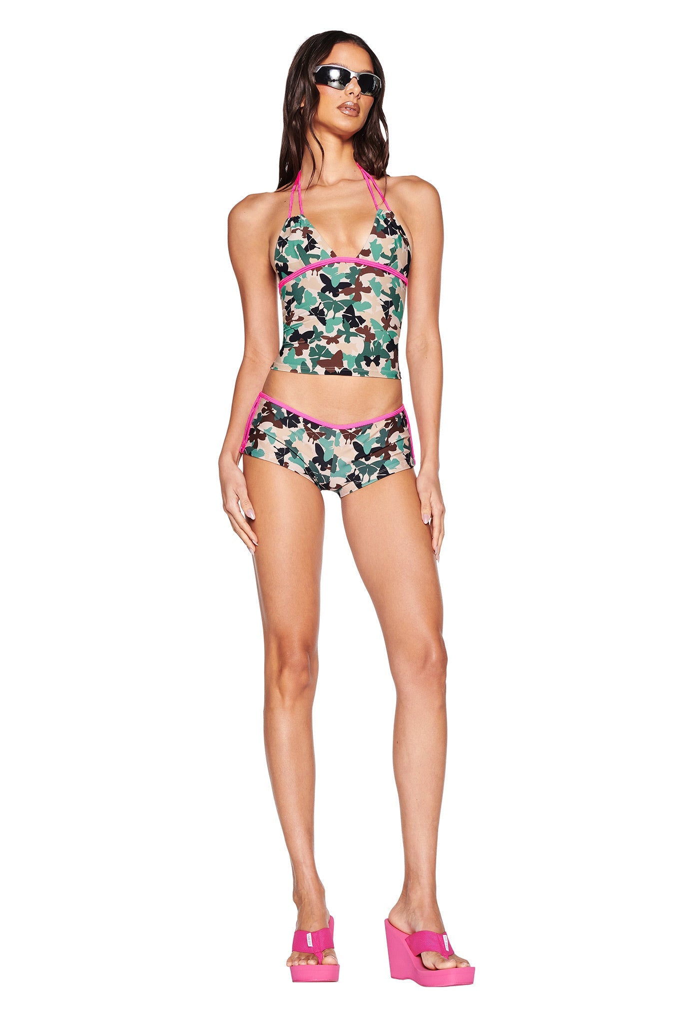 A woman wears JOANNE SHORT - FLUTTER CAMO by I.AM.GIA, featuring a green, pink, and beige pattern with pink trim. She pairs it with a matching bikini top, black sunglasses, and hot pink platform sandals while standing against a white background.