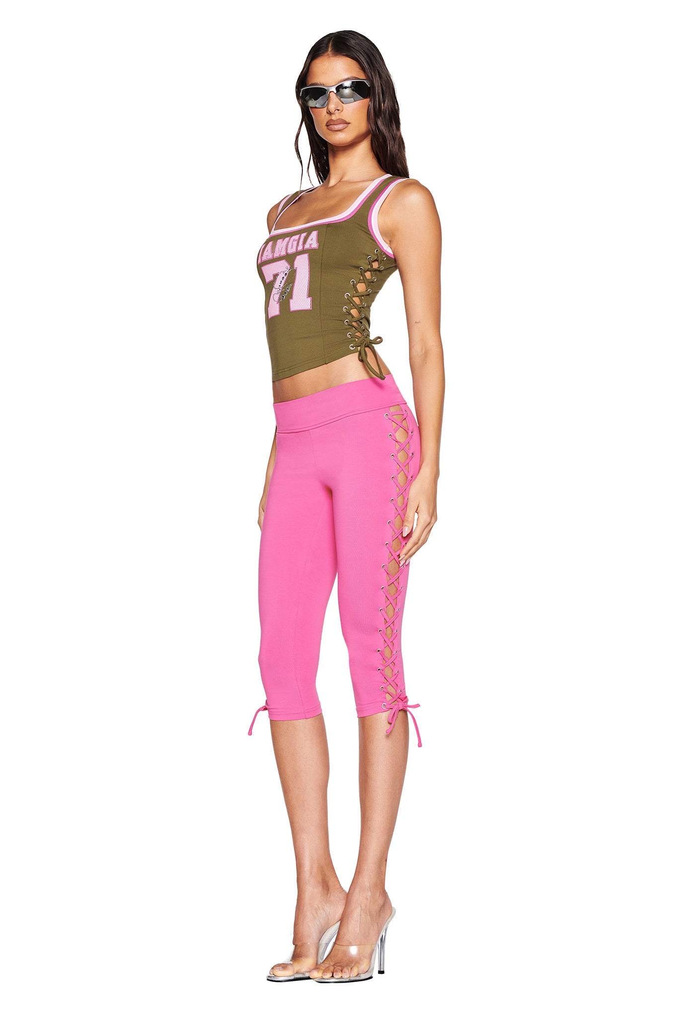 Someone sports sleek sunglasses, the "NOA TOP - GREEN" by I.AM.GIA, and pink capri pants with adjustable lace-up details. Clear high-heeled shoes complete the look against a plain white background.