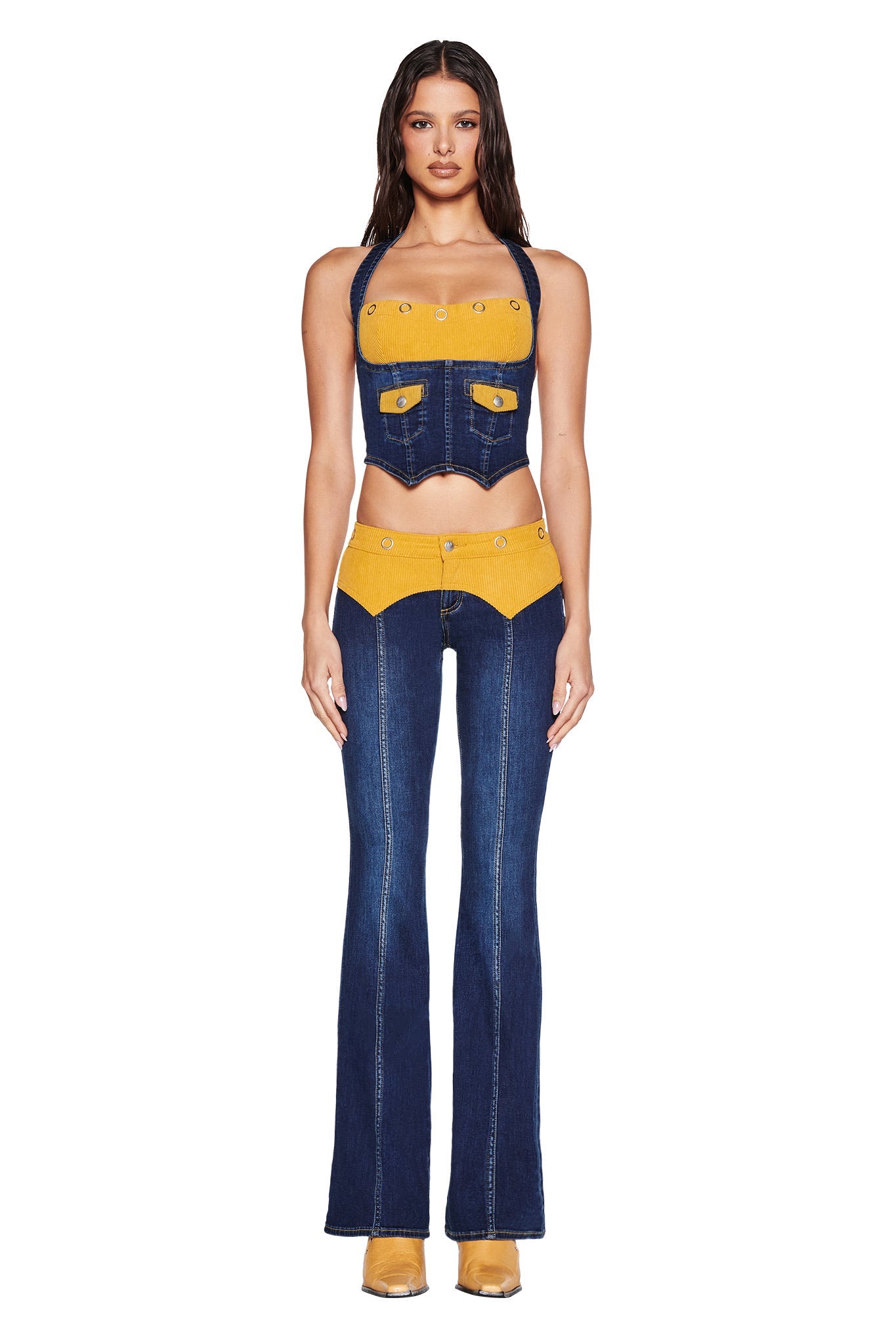 A woman faces forward in the I.AM.GIA TOMMIE CORSET - DARK WASH, a dark denim halter crop top with faux pockets, paired with matching high-waisted flared jeans featuring mustard corduroy details and tan boots. The background is plain white.