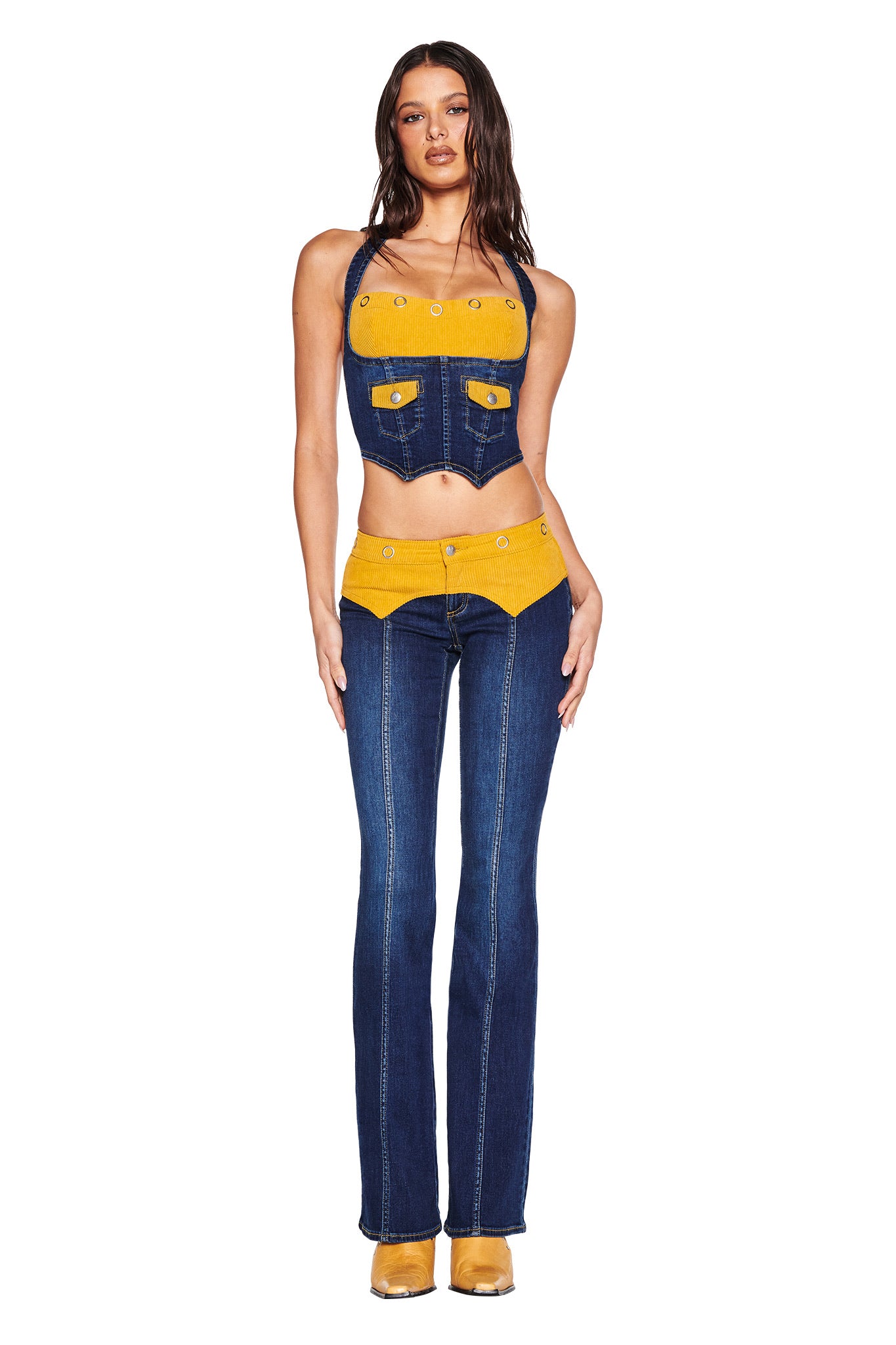 A woman faces forward in the I.AM.GIA TOMMIE CORSET - DARK WASH, a unique denim corset top with yellow accents, jacket-style pockets and eyelet details, paired with flared jeans featuring a yellow waistband and matching yellow shoes.