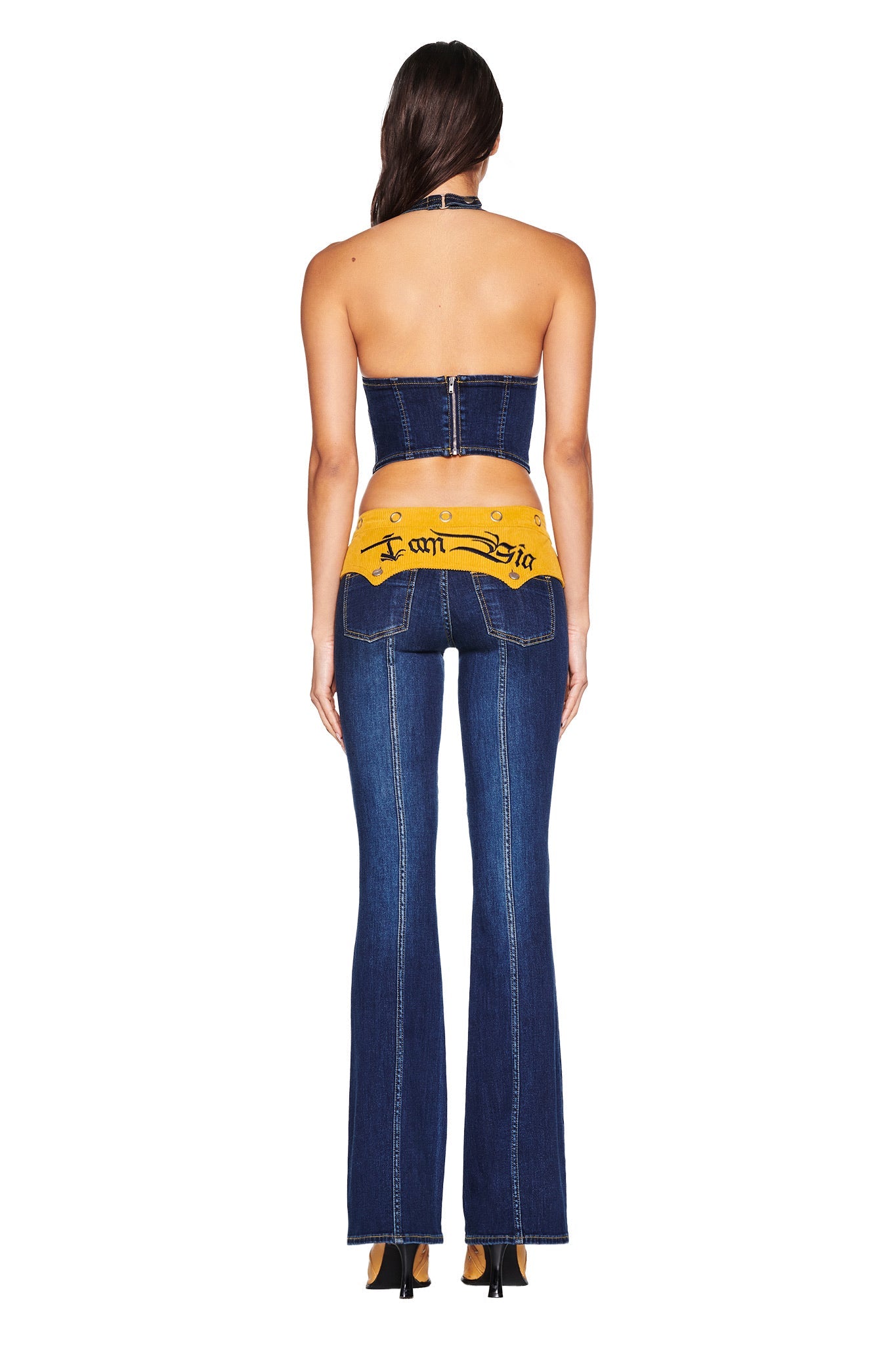 A woman faces away wearing the I.AM.GIA TOMMIE CORSET in dark wash, paired with flared denim jeans that have a yellow waistband with black gothic-style text, and finishes the look with black high heels.