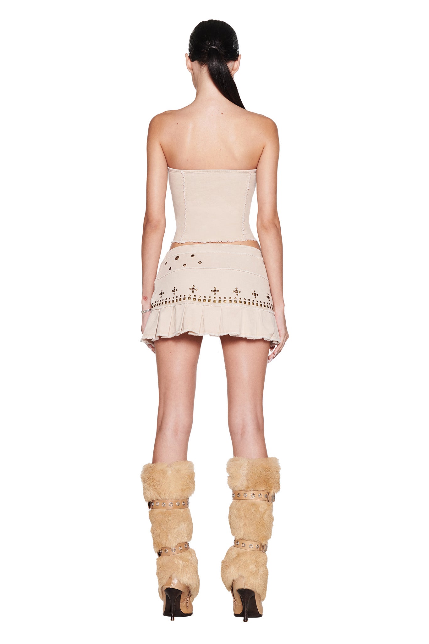 A woman with long dark hair in a ponytail faces away, wearing the I.AM.GIA MASON MINI SKIRT in tan—a canvas pleated mini skirt with an asymmetrical belt—styled with a strapless beige dress and tall tan fur boots.