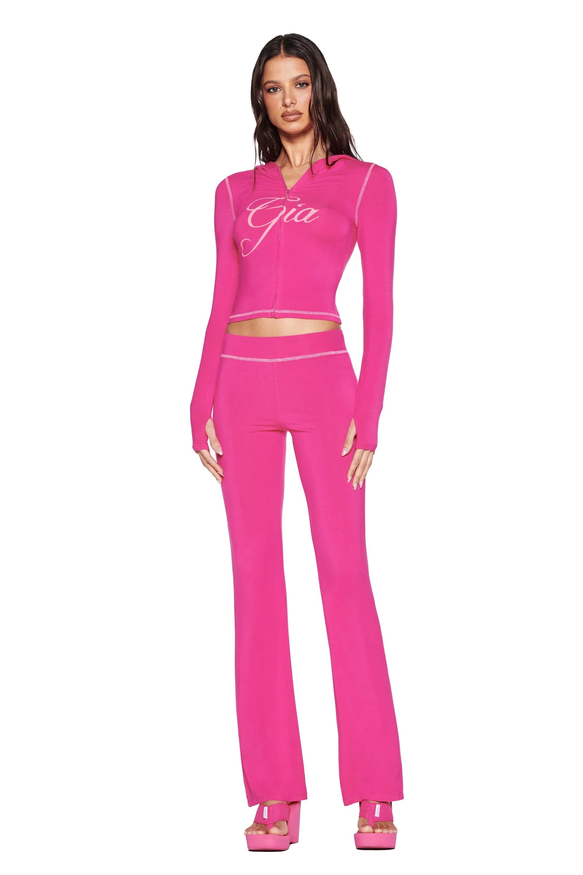 A woman poses against a white background in the I.AM.GIA BLARE TRACKPANT - HOT PINK with a matching long-sleeve logo top and platform heels, her wavy hair cascading down.