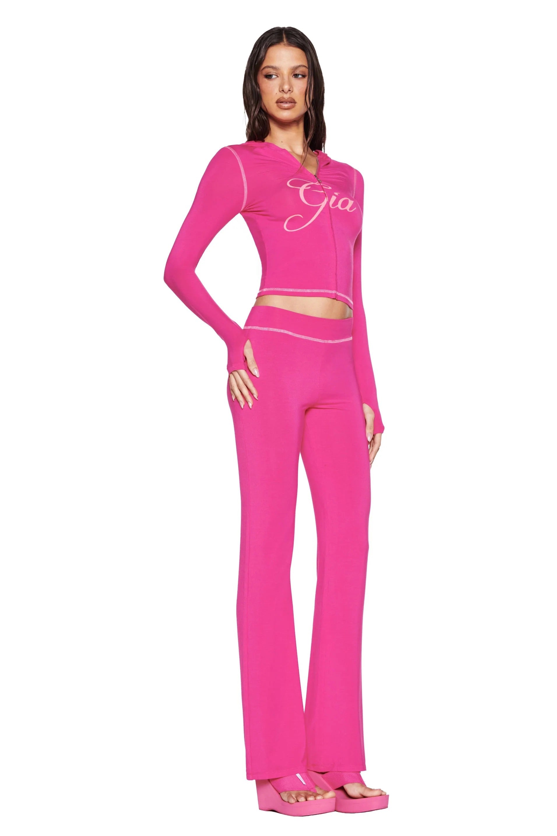 A woman with long dark hair poses against a white background wearing I.AM.GIA's hot pink BLARE TRACKPANT and matching zip-up top, paired with pink platform sandals.