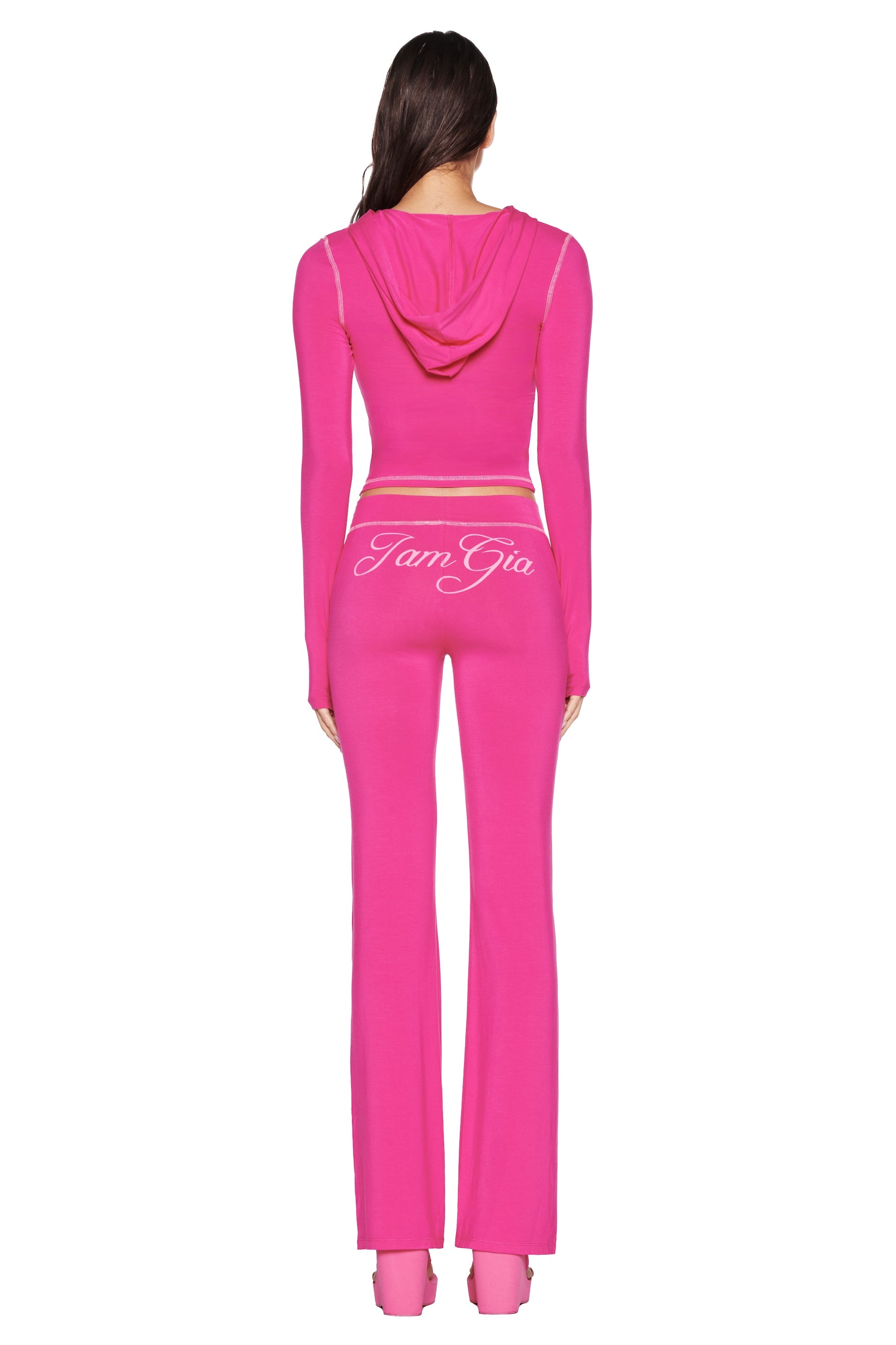 Seen from the back, a person with long, dark hair wears the I.AM.GIA BLARE TRACKPANT - HOT PINK outfit, featuring "Jam Gia" on the flared pants and a fitted jacket.