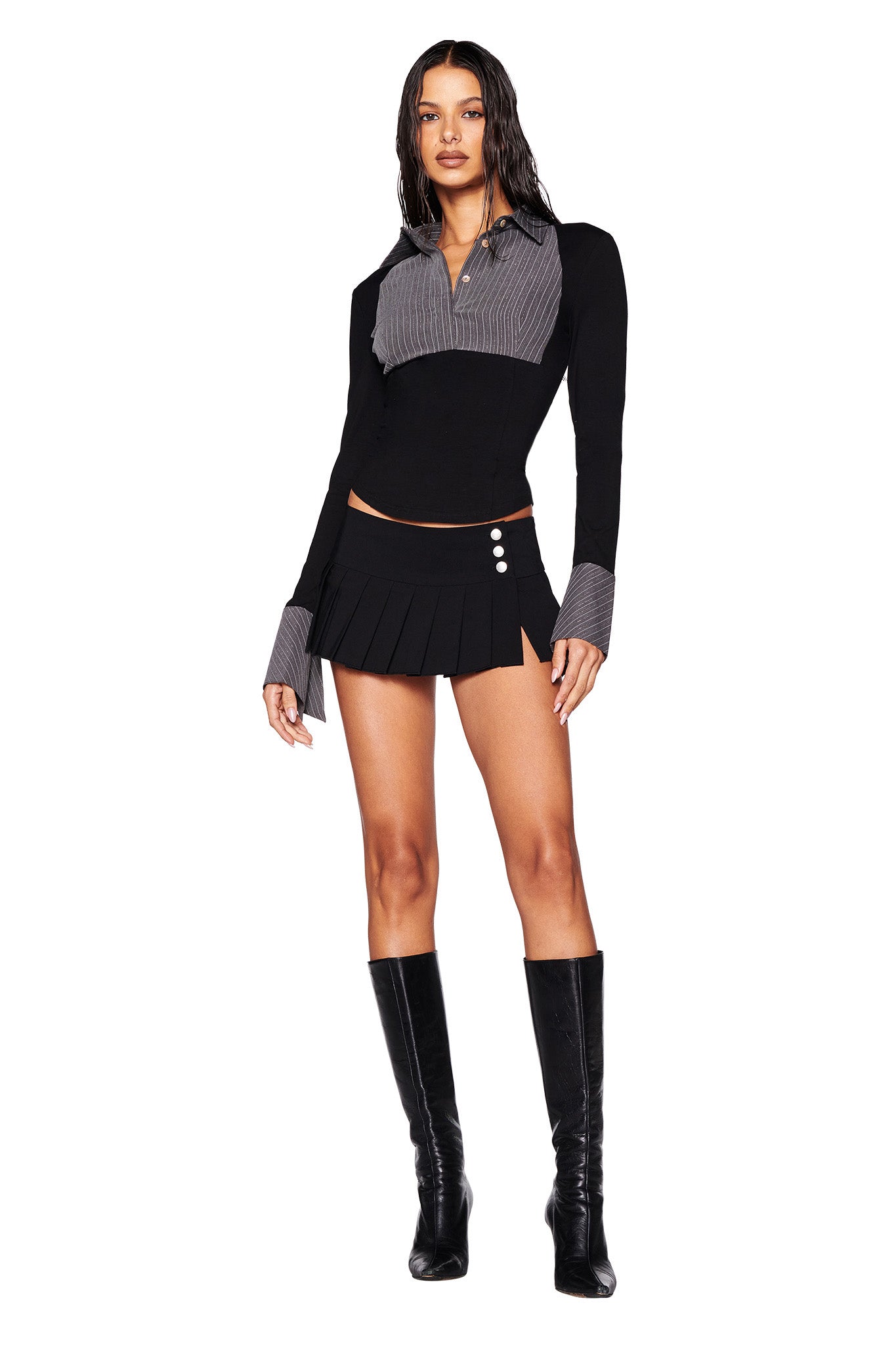 A woman stands wearing the SHARAH TOP in black and gray from I.AM.GIA, matched with a pleated mini skirt featuring button details and a bodycon fit. She completes her outfit with knee-high black boots. The background is white, and she has wet, wavy hair.