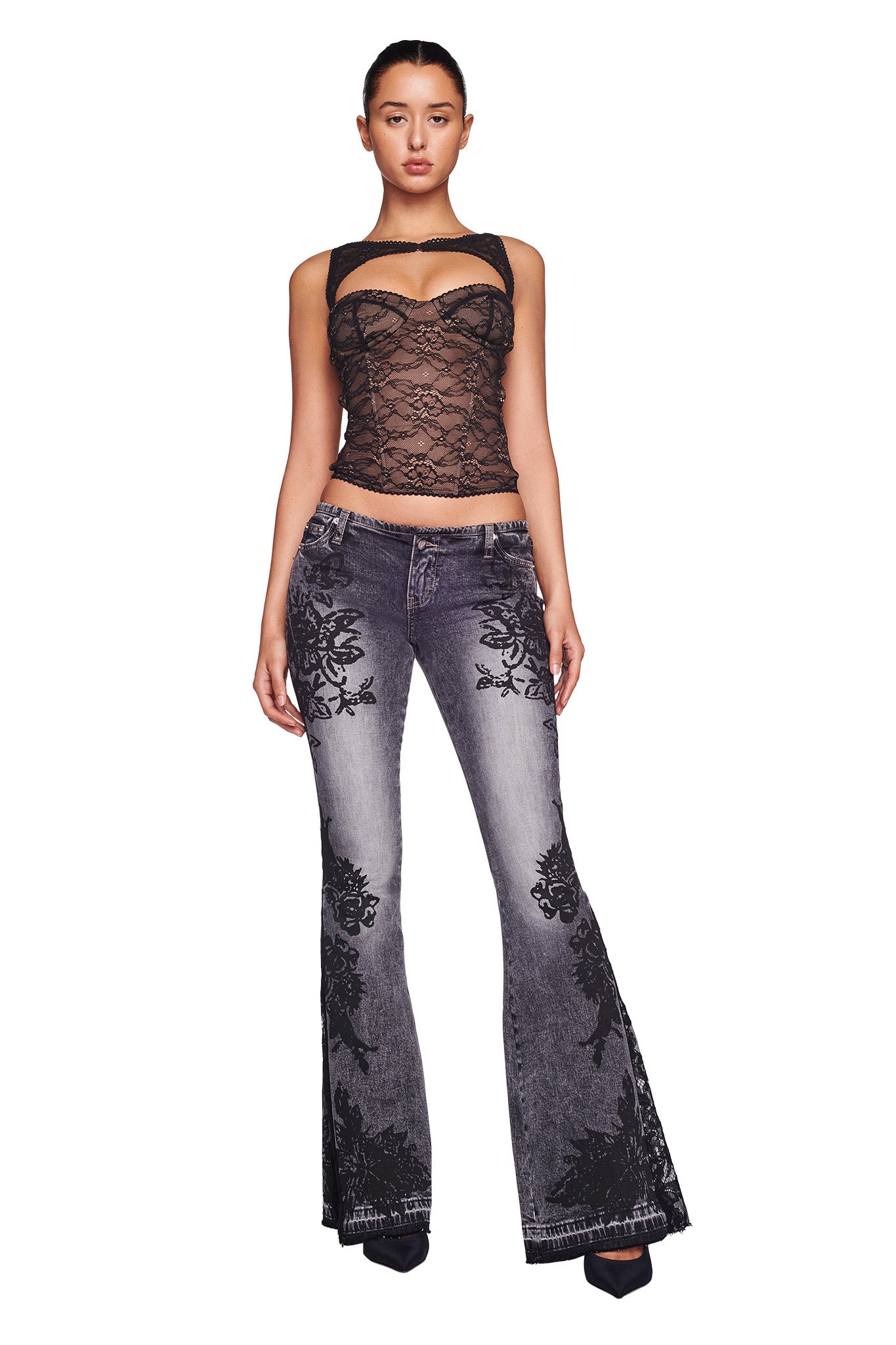 A woman stands against a white background, wearing the KIANA BUSTIER - BLACK from I.AM.GIA paired with grey flared jeans featuring black floral designs. Her hair is pulled back and she completes her look with black heeled shoes, gazing directly at the camera.