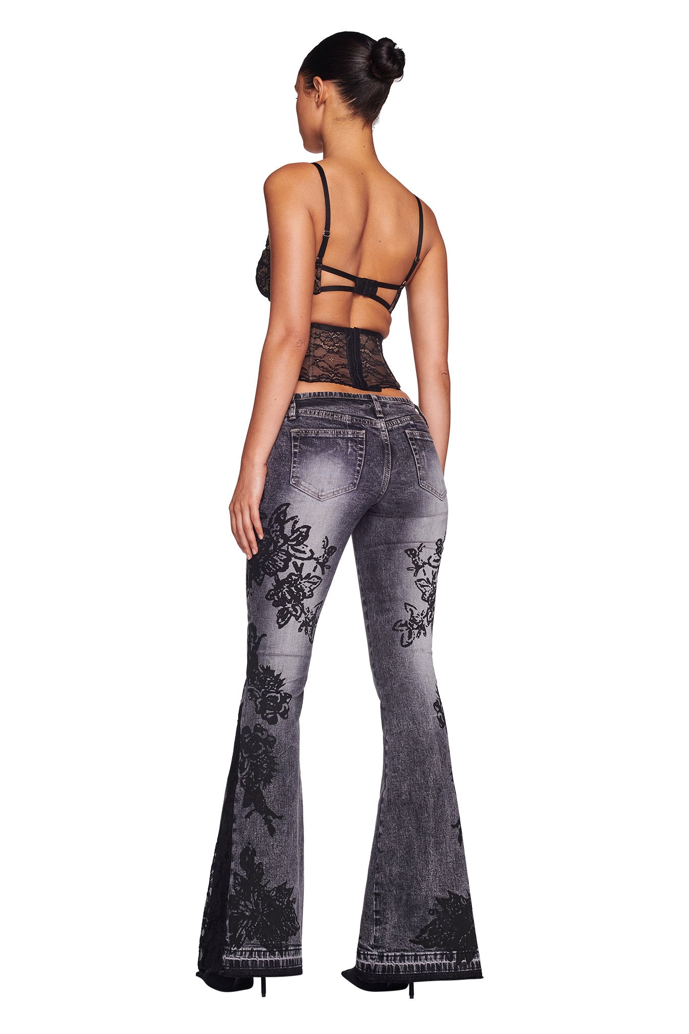 A woman stands facing away from the camera, wearing the KIANA BUSTIER - BLACK top and high-waisted, flared gray jeans with black floral designs from I.AM.GIA. She has tied-up hair and is wearing black high-heeled shoes. The background is plain white.