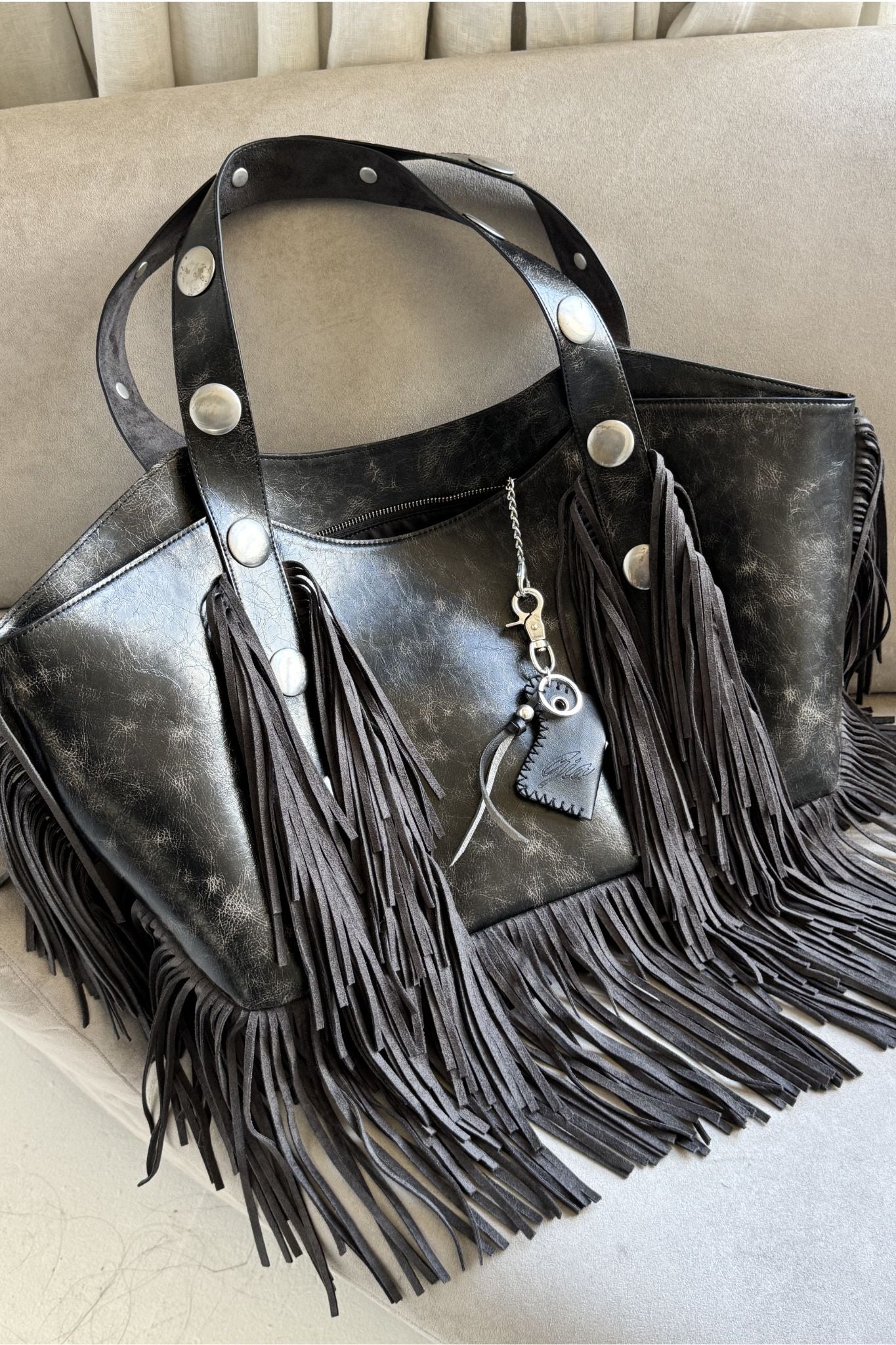The CHROMA FRINGE BAG - BLACK by I.AM.GIA, an oversized distressed tote with silver studs, long side and bottom fringe, and a leather heart keychain, rests elegantly on a beige surface.
