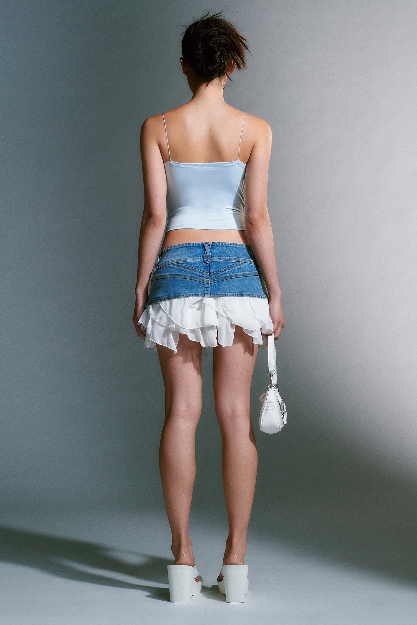 A person stands facing away, wearing an I.AM.GIA light blue satin camisole and a denim mini skirt with layered white ruffles. They hold a small white handbag in their left hand and wear I.AM.GIA SHELBY FLIP FLOP - WHITE sandals. The background is plain and softly lit.