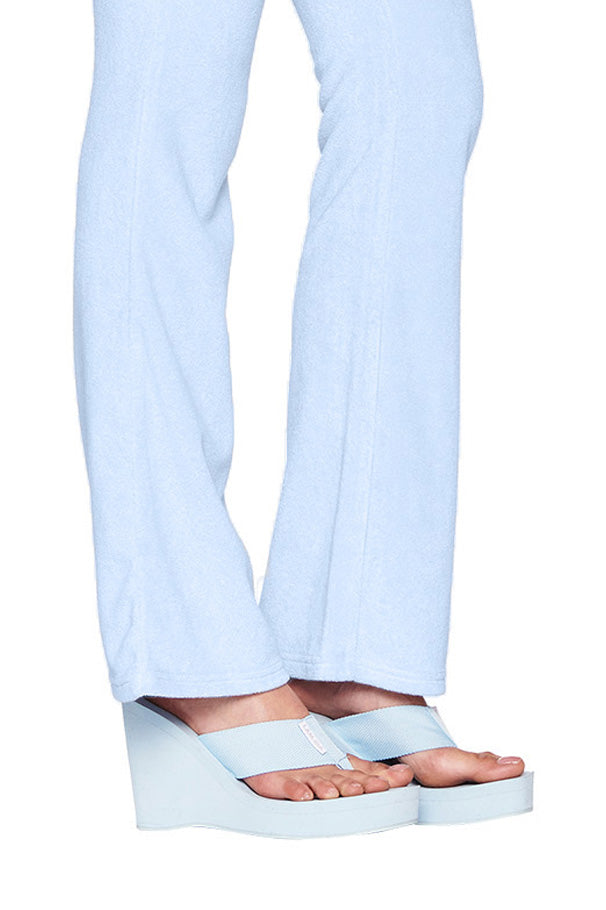 Close-up of a person's lower legs wearing light blue pants from I.AM.GIA and the SHELBY FLIP FLOP in BABY BLUE. The pants are flowing and slightly flared at the bottom, partially covering the shoes as the person stands. The background is plain white.