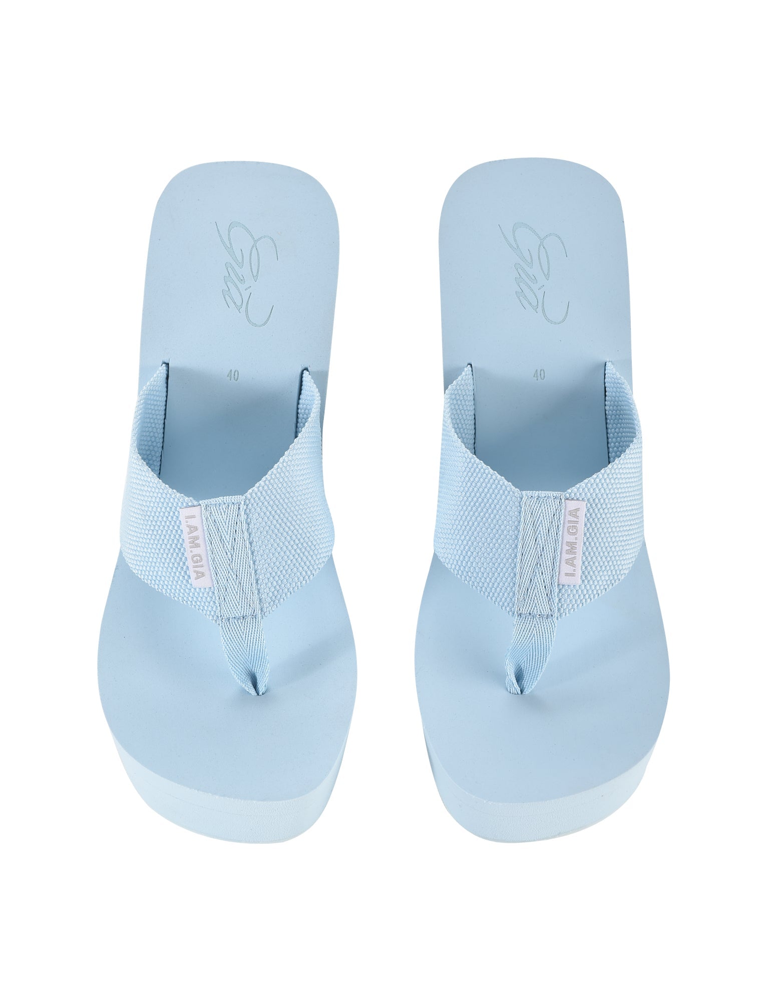 A pair of light blue flip-flops with wide, textured straps is displayed. The brand name "I.AM.GIA" is on a small tag on each strap, reminiscent of chic designs by the same brand. The SHELBY FLIP FLOP - BLUE : BABY BLUE flip-flops are placed side by side, showing the top view.