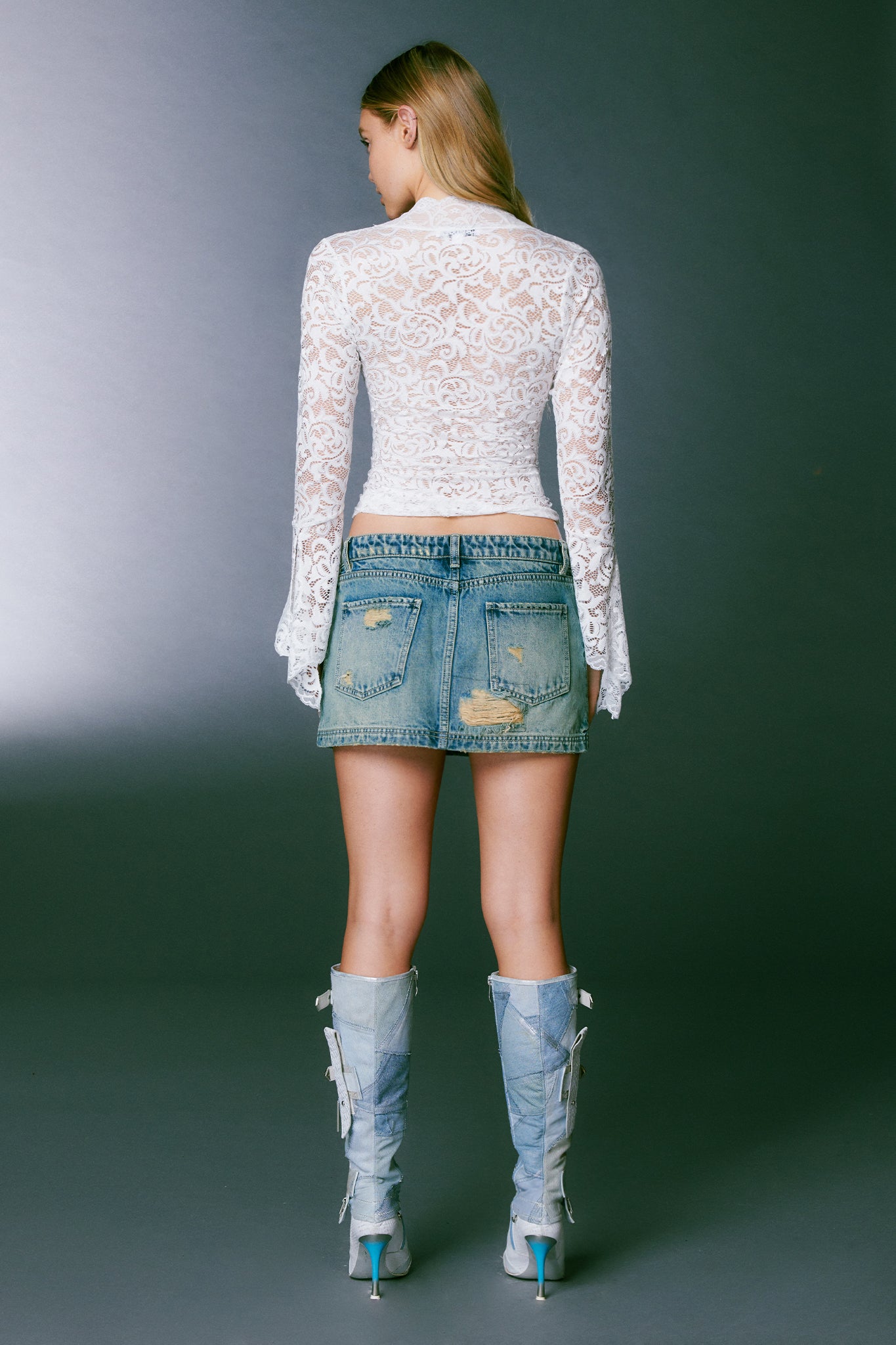 A woman stands facing away, showcasing the back of her outfit. She is wearing the GEMMA TOP - WHITE, a long-sleeved lace top from I.AM.GIA, paired with a short distressed denim skirt and knee-high blue boots with high heels. The background is a plain, dark gray.