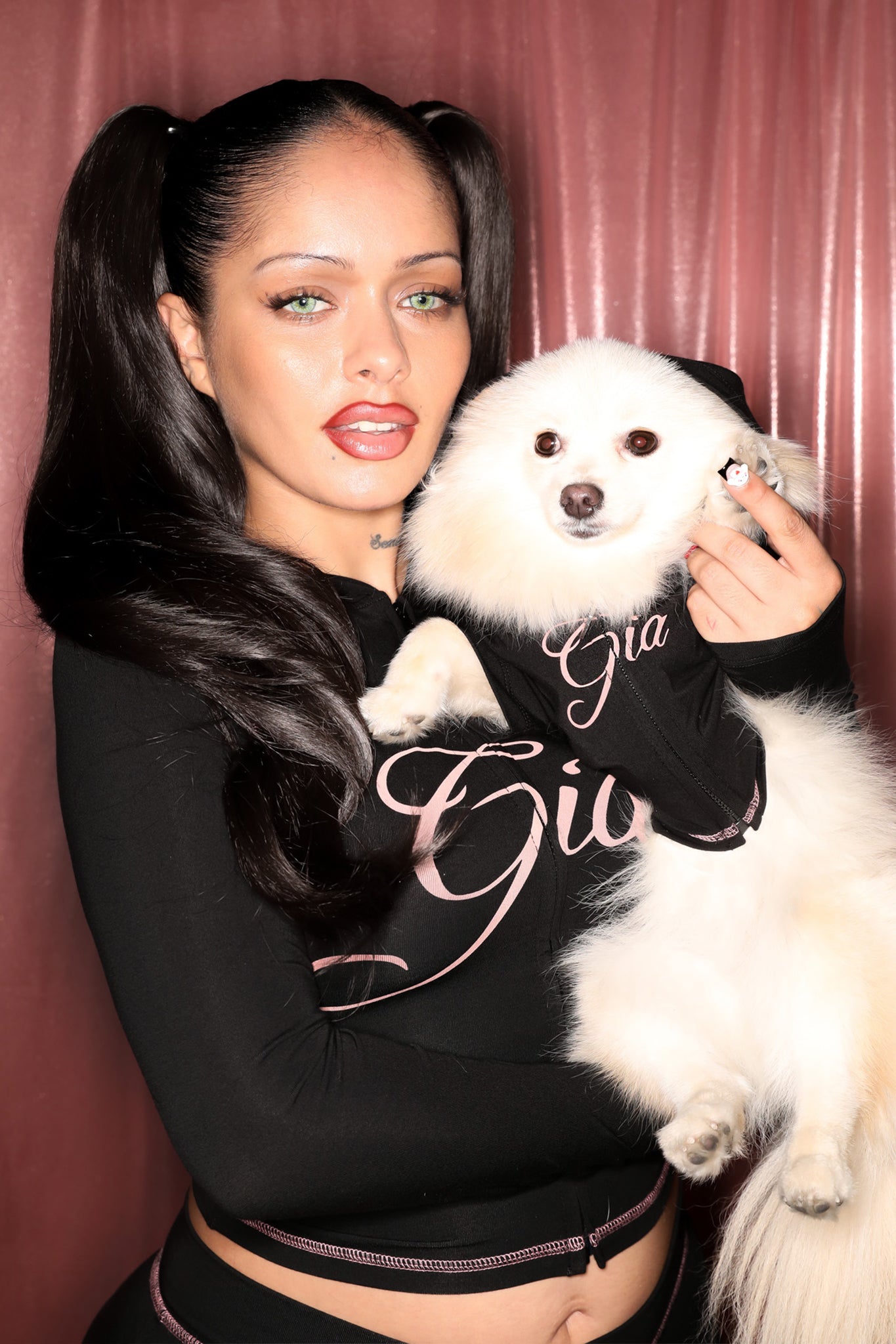A woman with long dark hair in pigtails and green eyes holds a fluffy white dog; both wear black outfits with pink writing, including the I.AM.GIA BLARE BEST FRIEND DOG HOODIE in black, standing in front of a pinkish curtain.