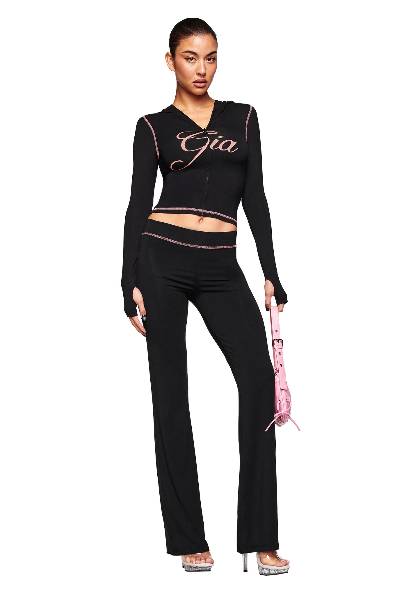 A woman stands confidently in I.AM.GIA’s black BLARE TRACKPANT and a matching fitted crop top with "Gia" lettering, holding a pink handbag and wearing silver platform heels against a clean white background.