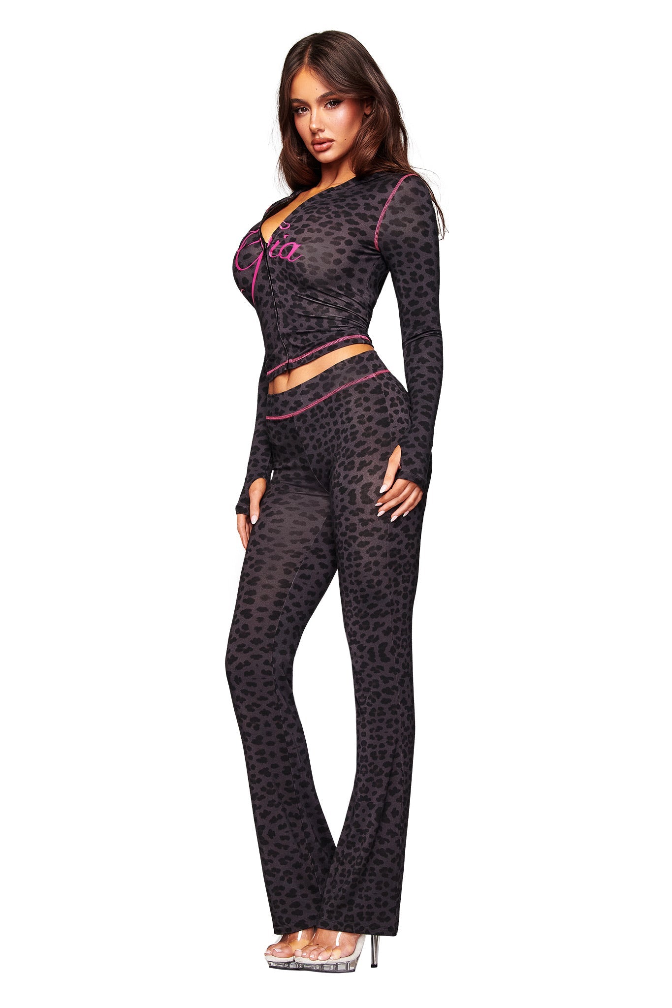 A woman models the I.AM.GIA BLARE TRACKPANT - PANTHER, a fitted, long-sleeve black and dark gray panther print outfit with pink accents, paired with clear high-heeled sandals against a plain white background.