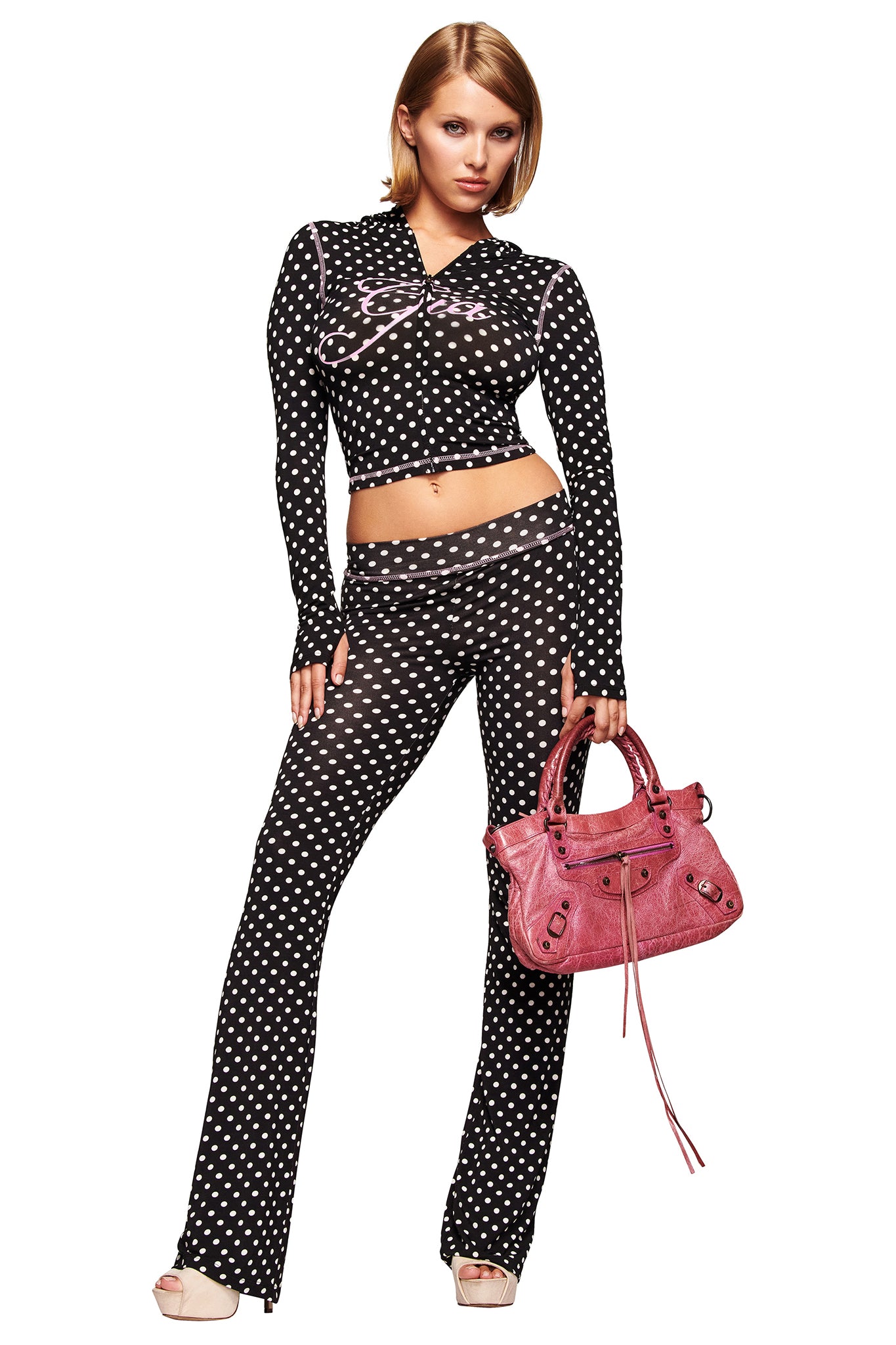 A woman poses against a white background wearing the I.AM.GIA BLARE HOODIE - BLACK POLKA DOT with matching flared pants, light-colored shoes, and holding a large pink handbag. Both hoodie and pants feature bold white polka dots.