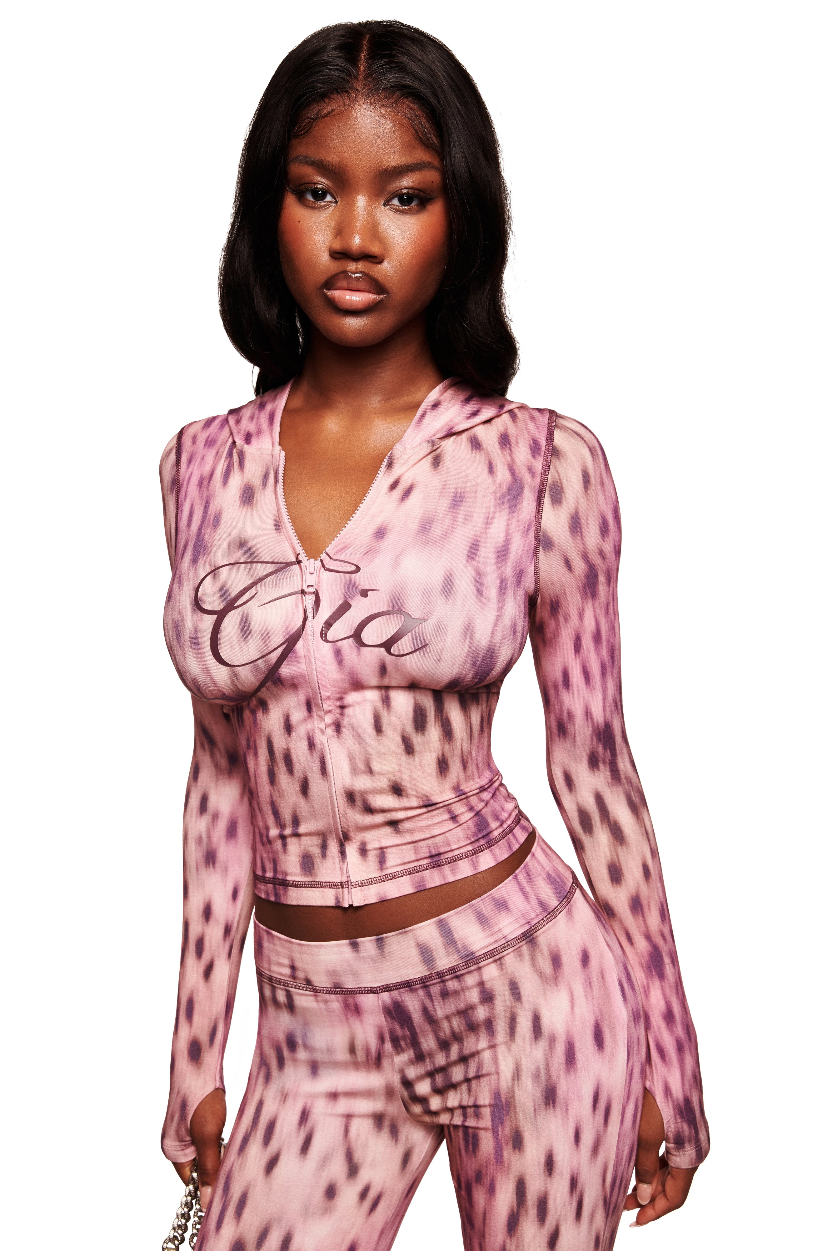 A woman wearing the I.AM.GIA BLARE HOODIE in pink blurred cheetah with "Gia" on the chest, matching high-waisted pants, and holding a small chain strap purse stands against a white background.