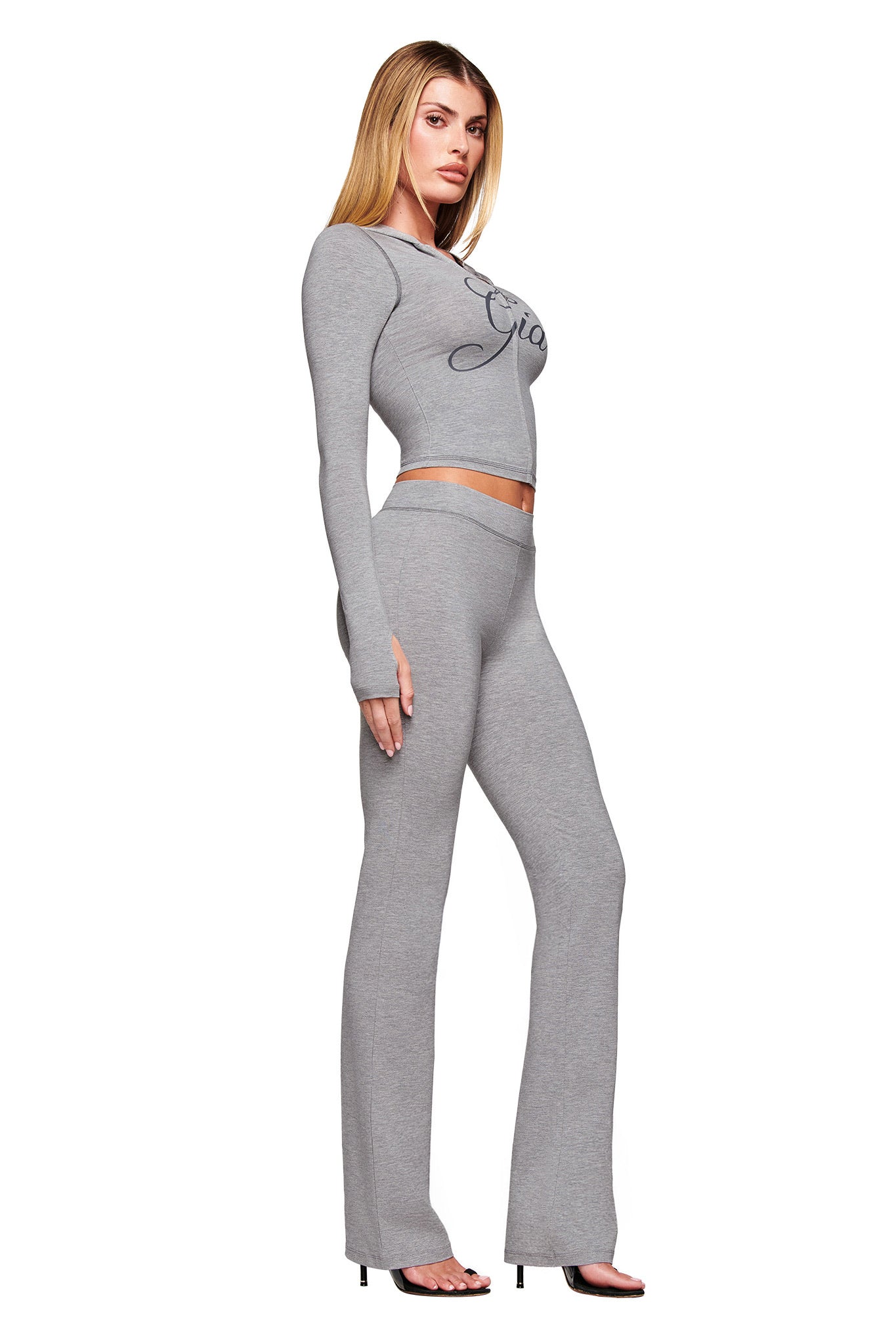 A woman poses in a fitted, heather grey, long-sleeve crop top and matching I.AM.GIA BLARE JERSEY TRACKPANT - GREY with flared legs. She wears black open-toe heels and has straight blonde hair against a white background.