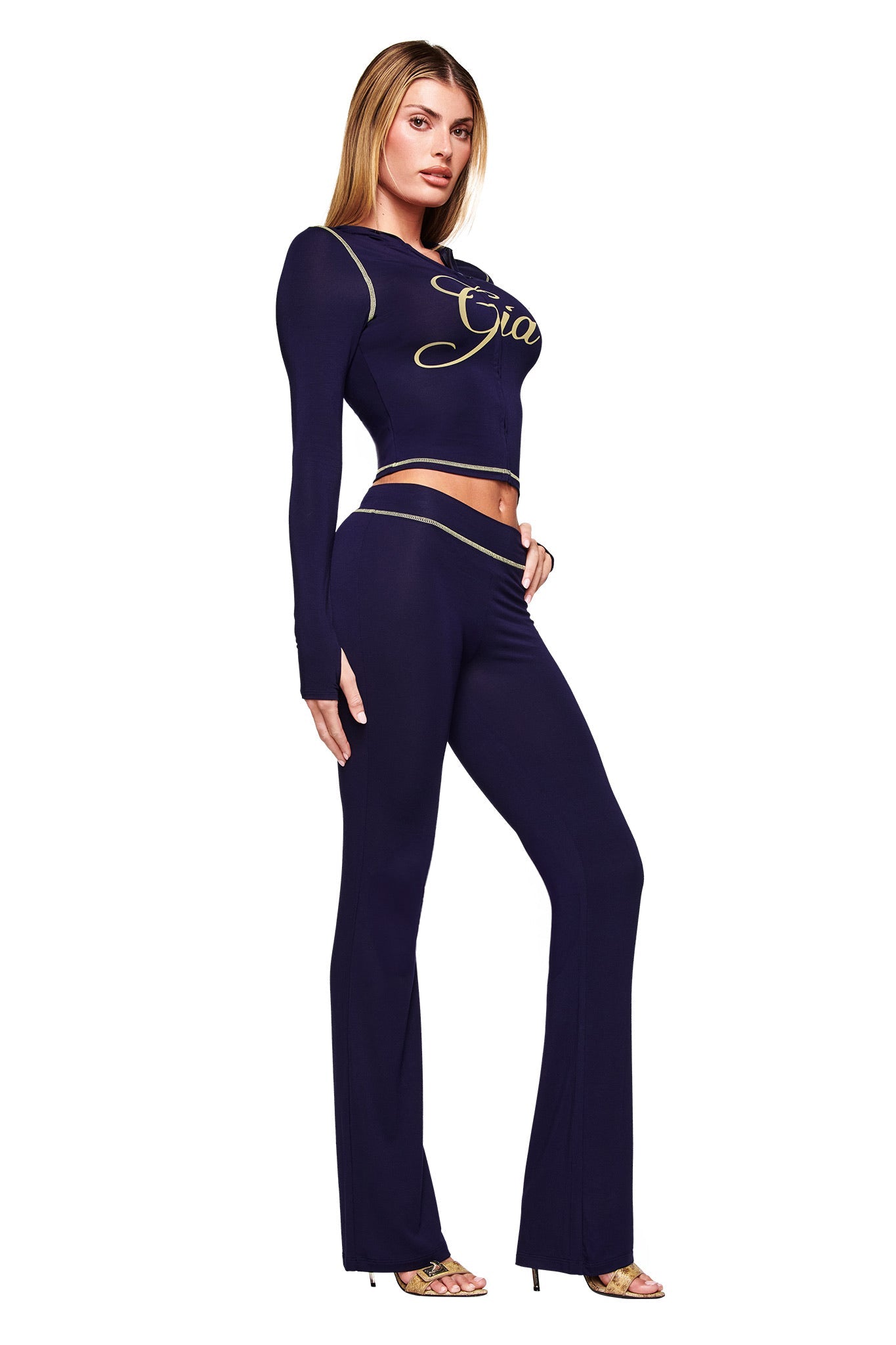A woman faces slightly sideways, wearing the I.AM.GIA BLARE HOODIE in navy with gold lettering and matching high-waisted flared pants. She pairs it with heels, her straight light brown hair down, against a white background.