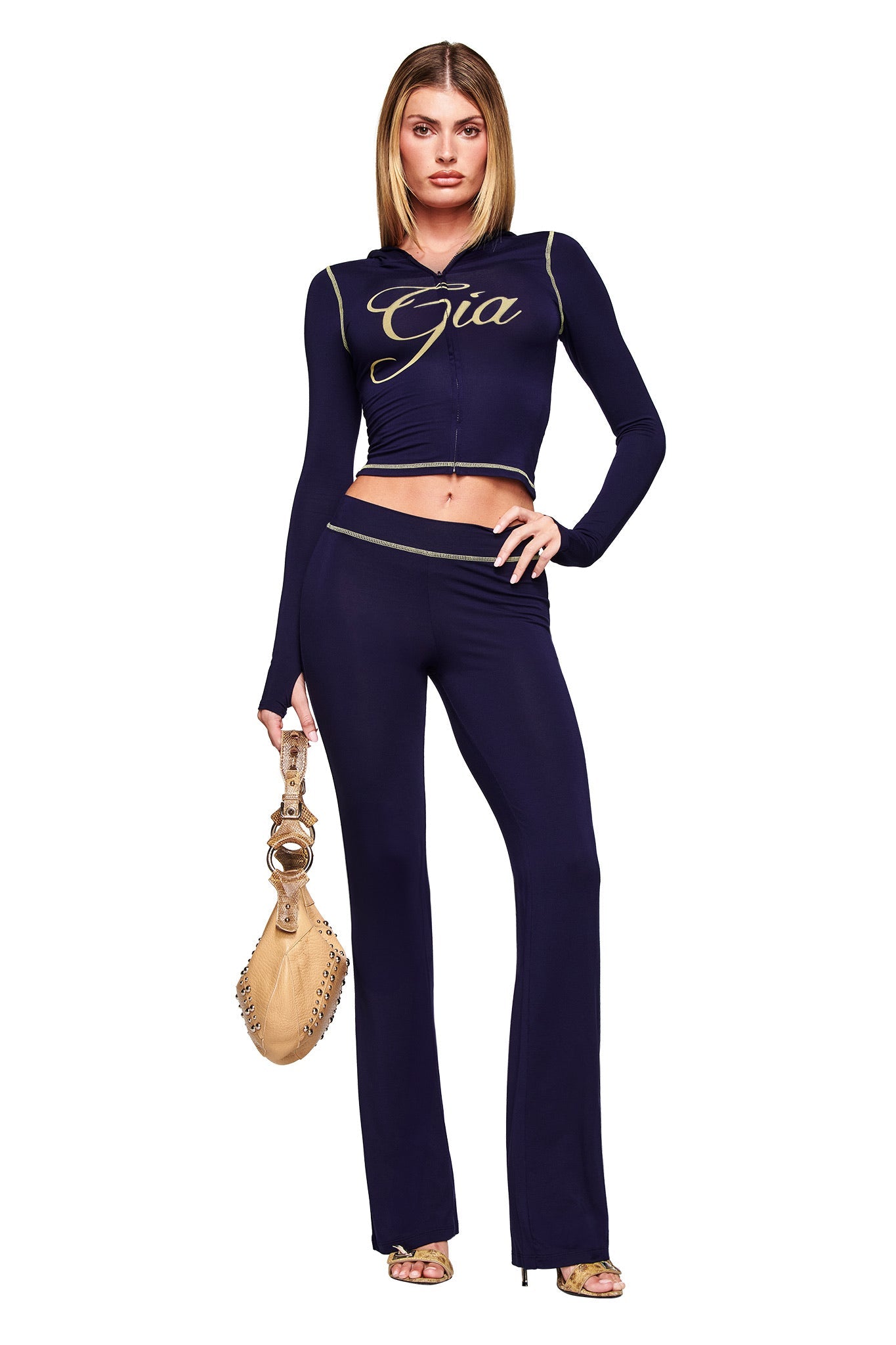 A woman stands confidently in I.AM.GIA's BLARE TRACKPANT - NAVY, paired with a matching long-sleeve crop top featuring “Gia” in gold script. She accessorizes with a tan handbag and gold heels.