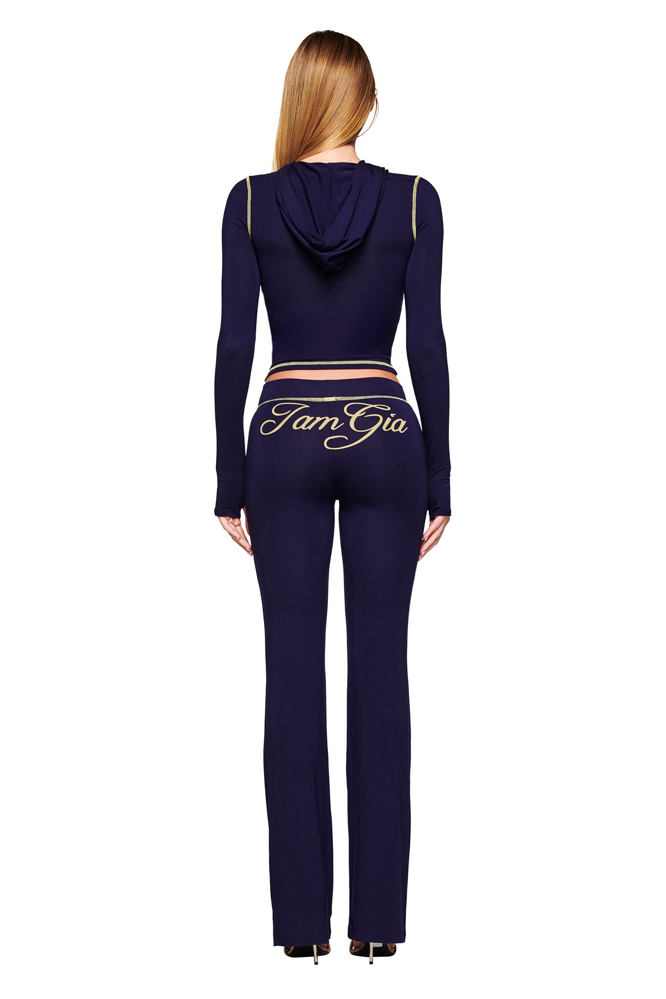A woman faces away, wearing I.AM.GIA's BLARE TRACKPANT in navy with gold accents and "I.AM.GIA" script on the back. The flared leg pants pair with a matching hooded top, complementing her long straight hair and heels.