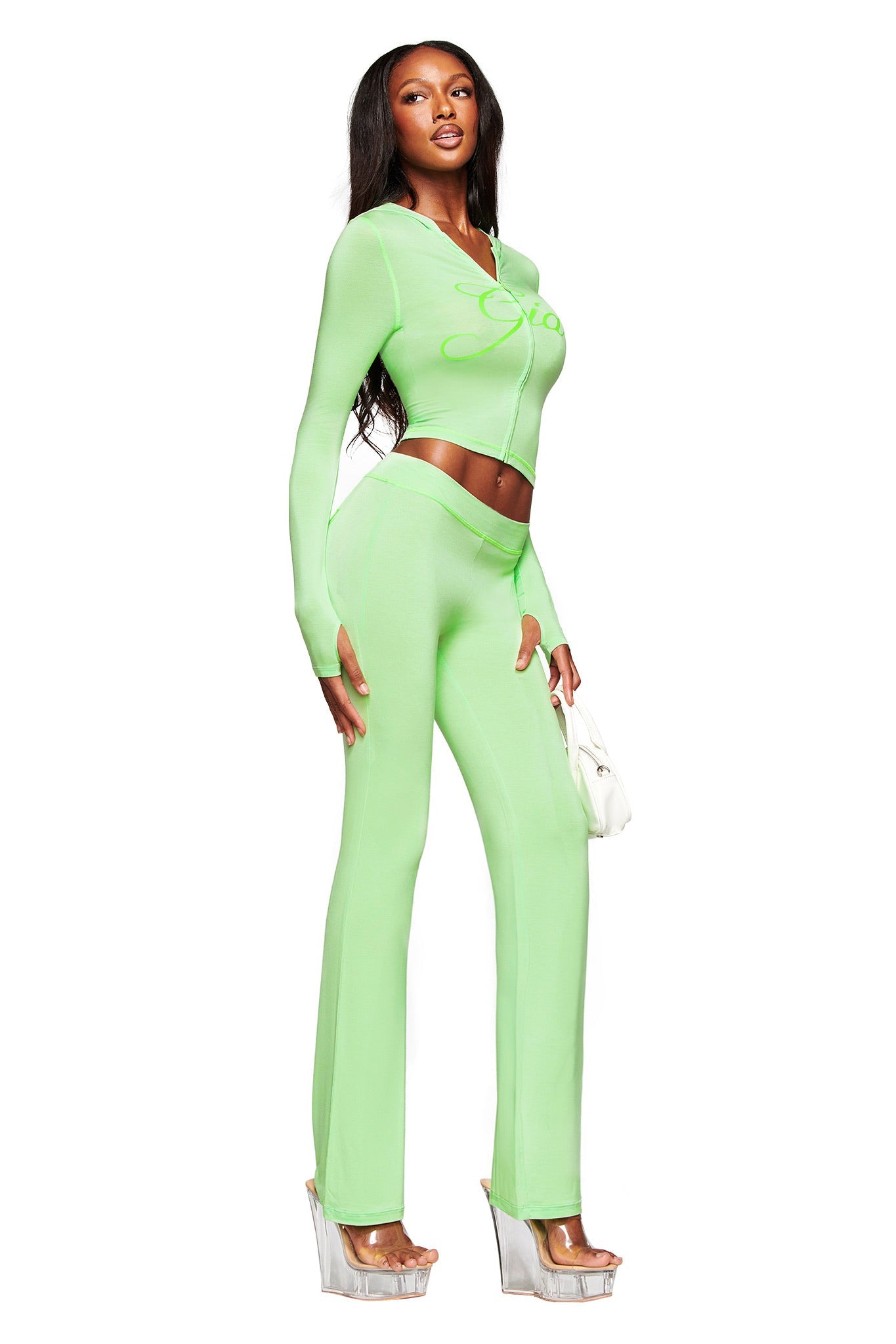 A woman wears the I.AM.GIA BLARE HOODIE - NEON GREEN two-piece set with long sleeves and flared pants, holding a small white handbag and wearing clear platform heels, standing against a white background.
