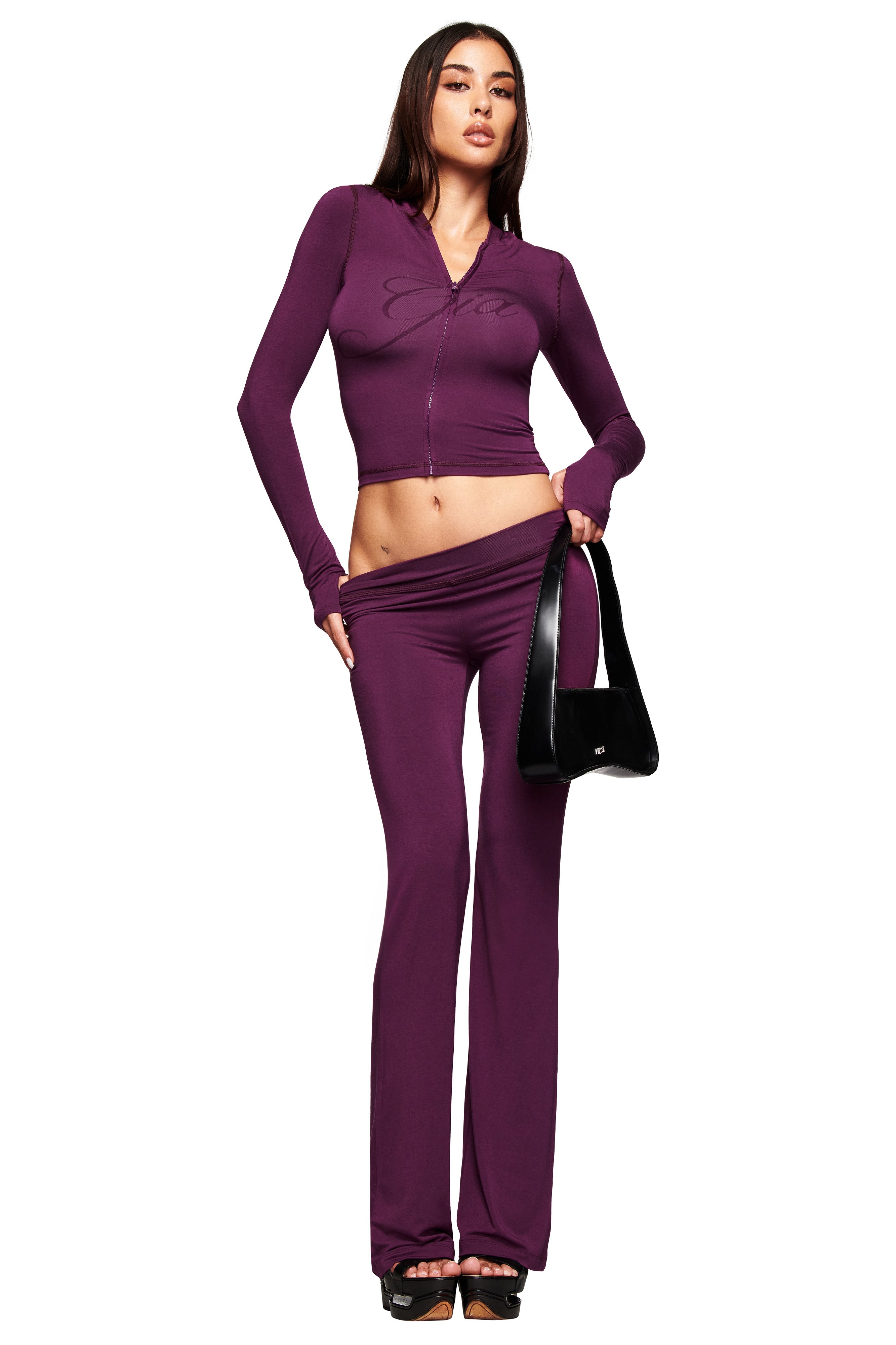 A woman models the I.AM.GIA BLARE TRACKPANT in plum with a matching fitted zip-up top, accessorized with a black handbag and platform sandals against a white background.