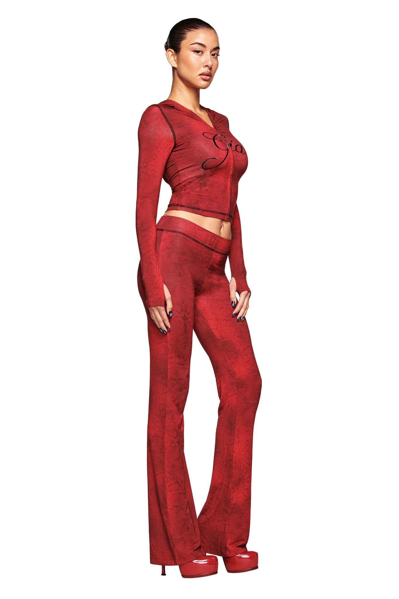 A woman poses against a white background in I.AM.GIA’s BLARE TRACKPANT - RED SERPENTINE and a matching fitted long-sleeve top, styled with platform heels.