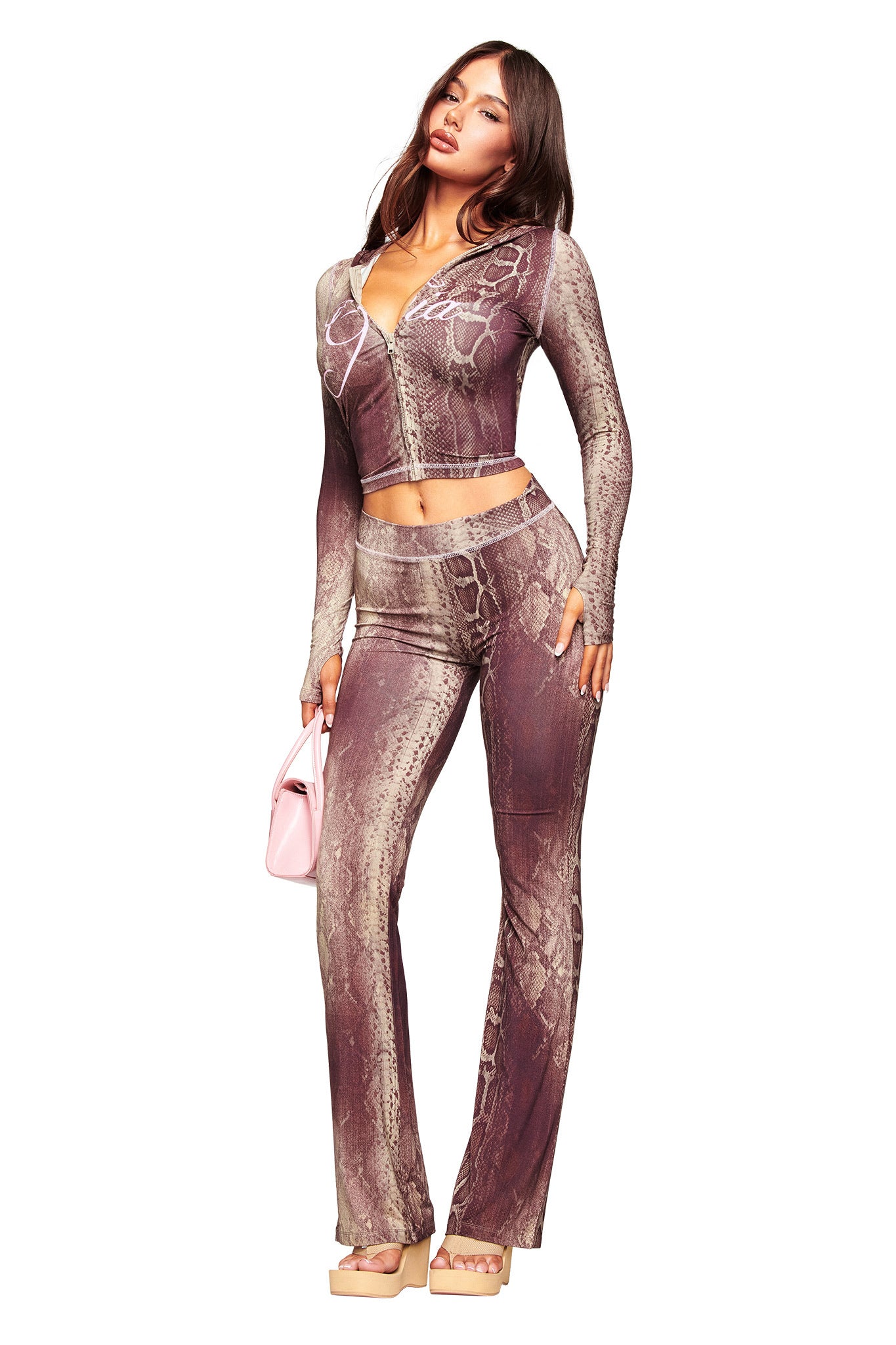 A woman poses in a fitted, long-sleeve snake print top and I.AM.GIA BLARE TRACKPANT - SNAKE, featuring flared legs. She holds a small pink handbag and wears nude platform sandals against a white background.