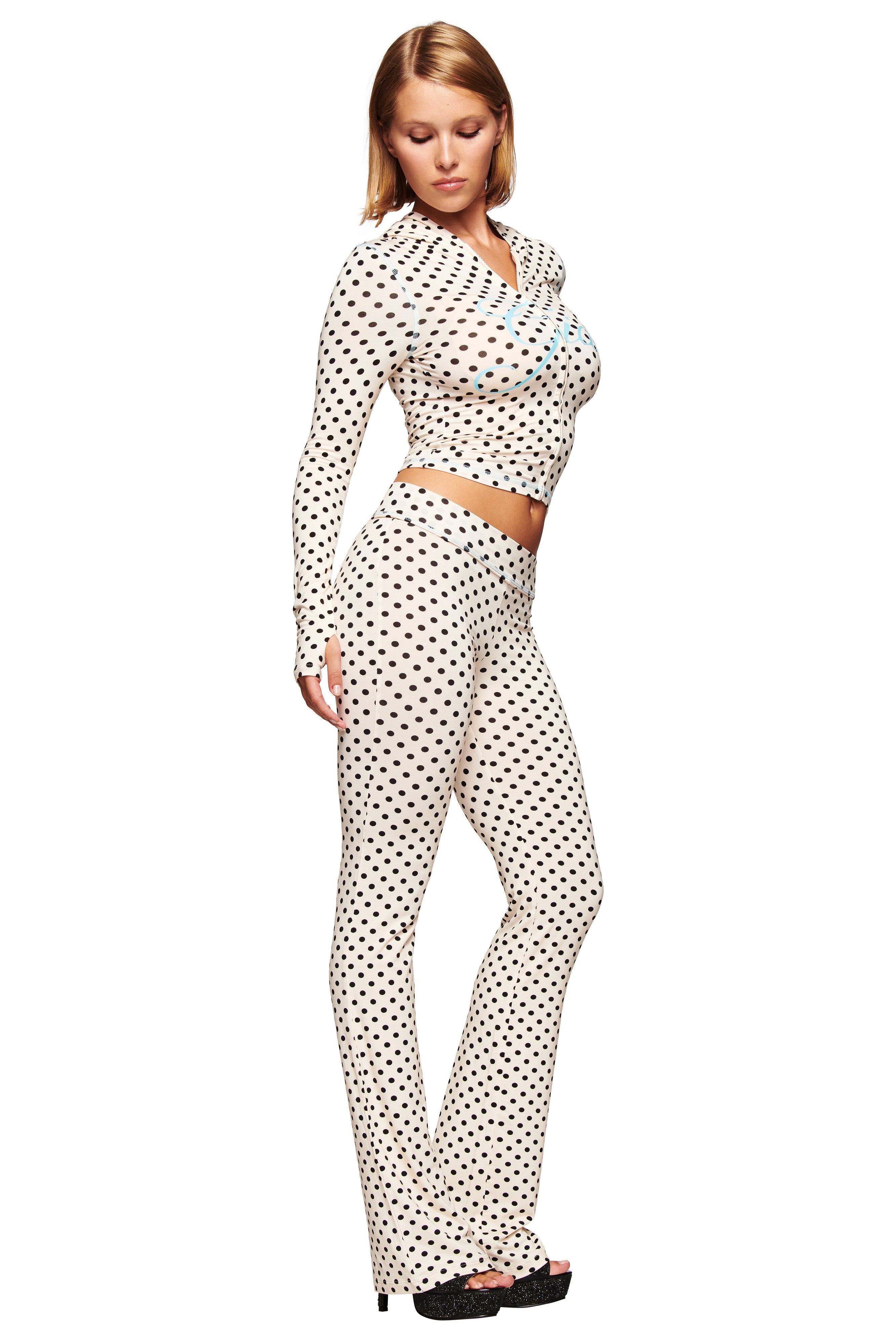 A woman stands in a neutral pose, looking down, wearing the I.AM.GIA BLARE TRACKPANT in cream polka dot—a fitted, long-sleeve outfit with flared legs—against a plain white background.