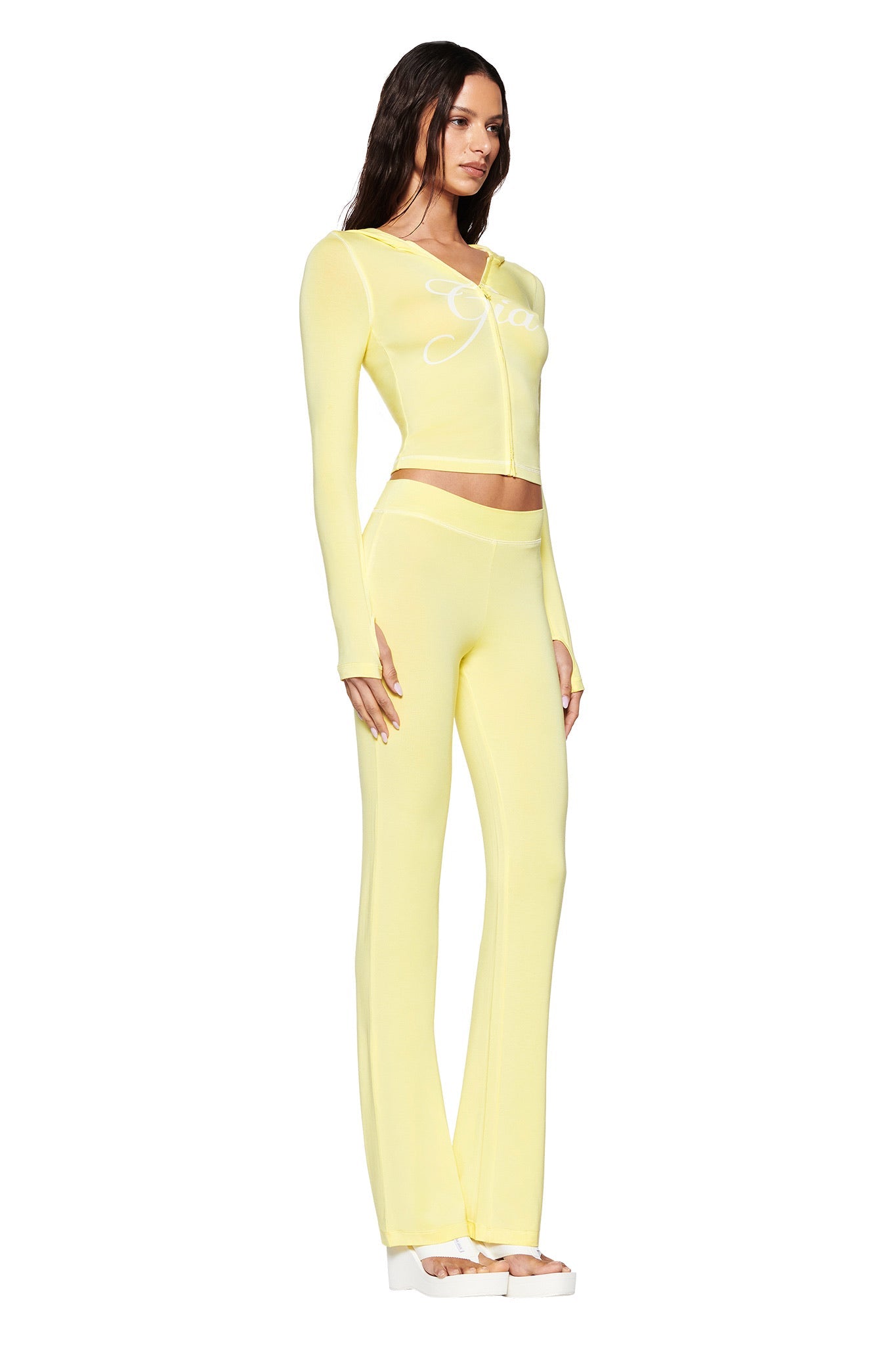 A woman in a light yellow outfit flaunts an I.AM.GIA Bamboo Jersey Long-Sleeve Top with "Gradient" text and matching high-waisted Blare Trackpant—Yellow while wearing white platform shoes, her style subtly accentuated against a plain white background.